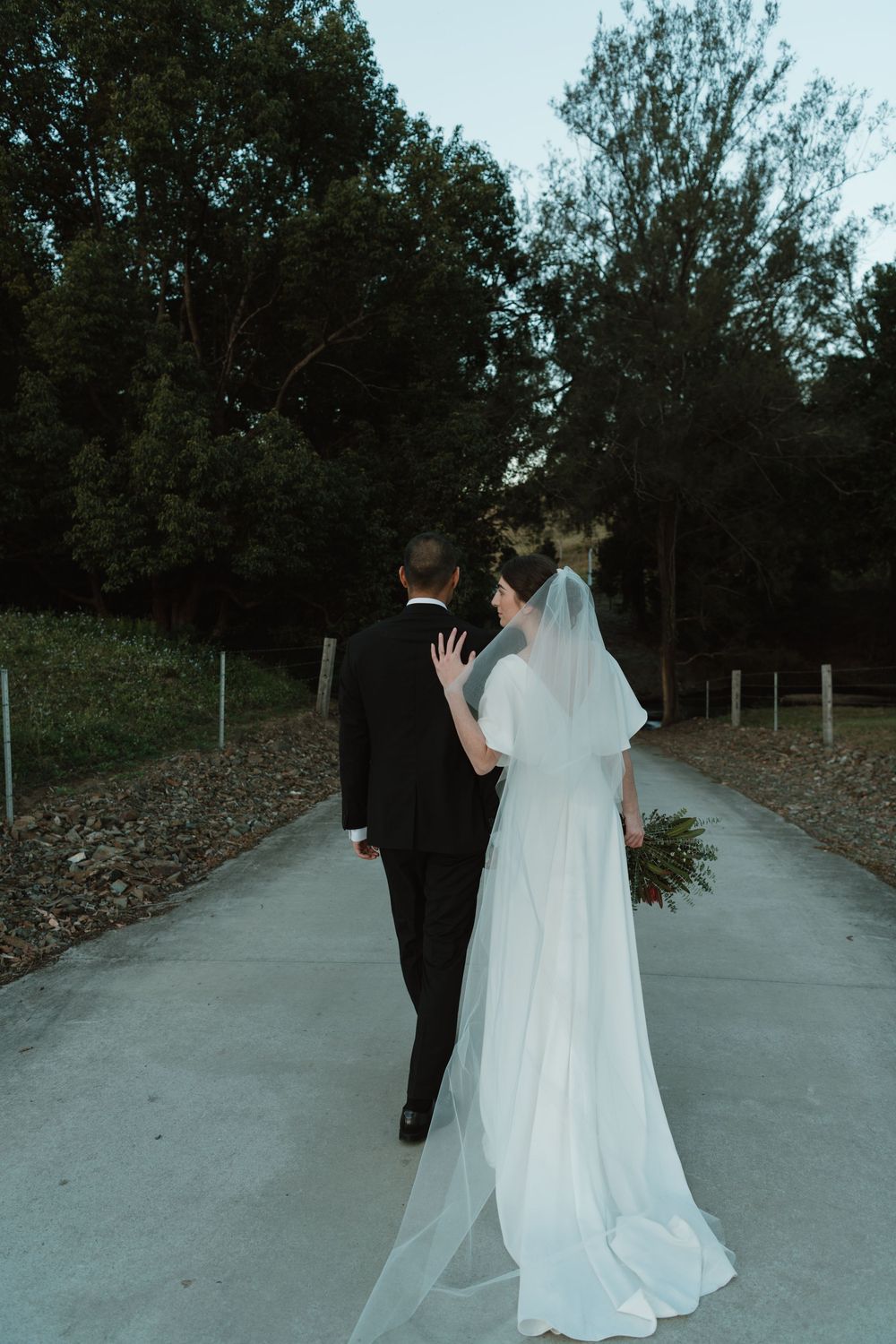 Bride and groom walking hand in hand down a path surrounded by lush green trees on their wedding day.