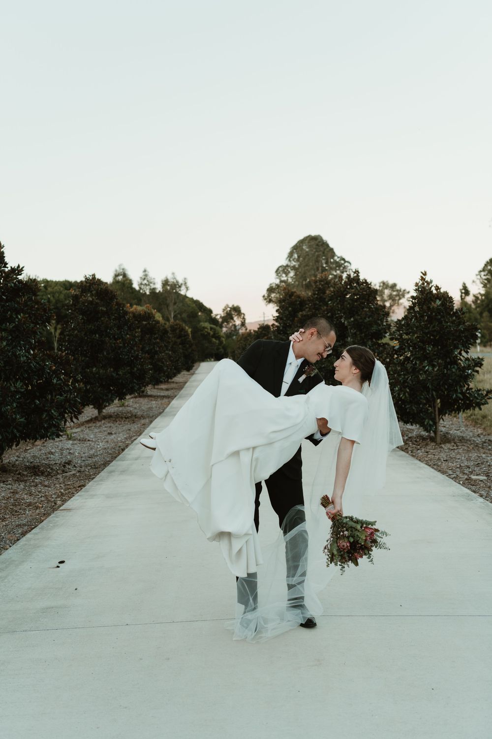 Groom lifts bride in white dress and boots while sharing a kiss on a pathway surrounded by lush green trees.