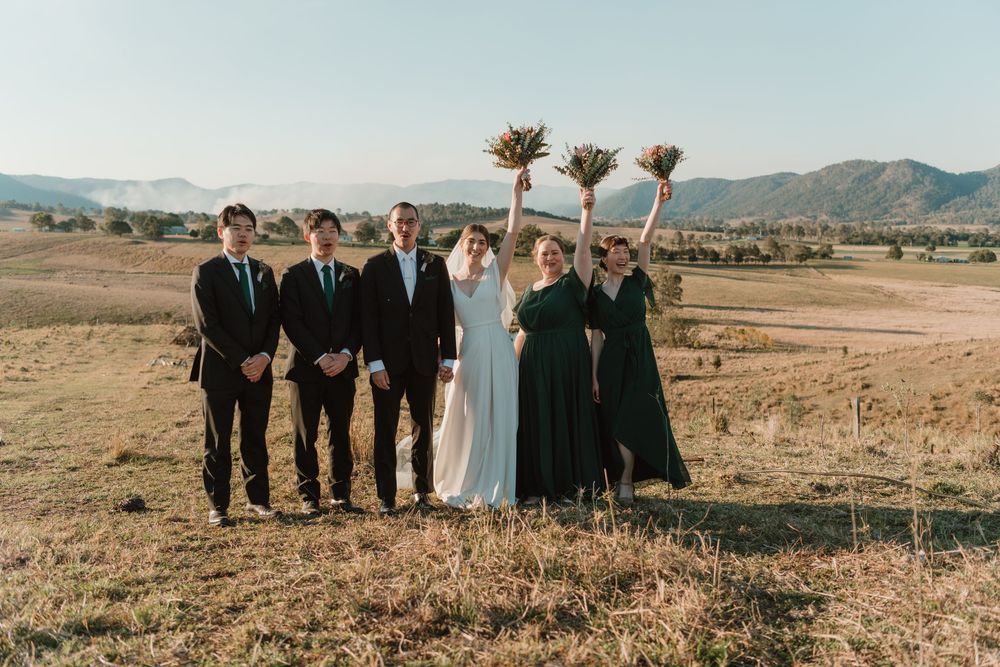 Wedding party poses outdoors with rolling hills backdrop, bride in white, bridesmaids in green raising bouquets.