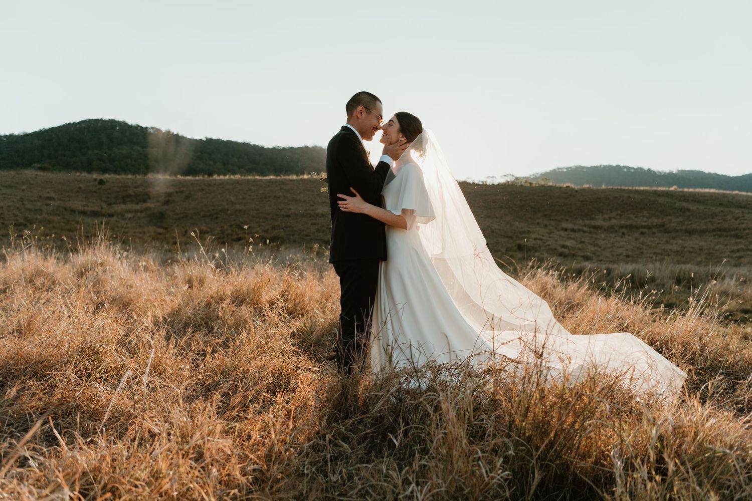 Bride and groom share a romantic kiss in a golden field with rolling hills, wearing elegant wedding attire.