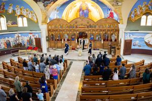 Interior view of an ornate Orthodox church with wooden pews and golden iconostasis during a religious ceremony.