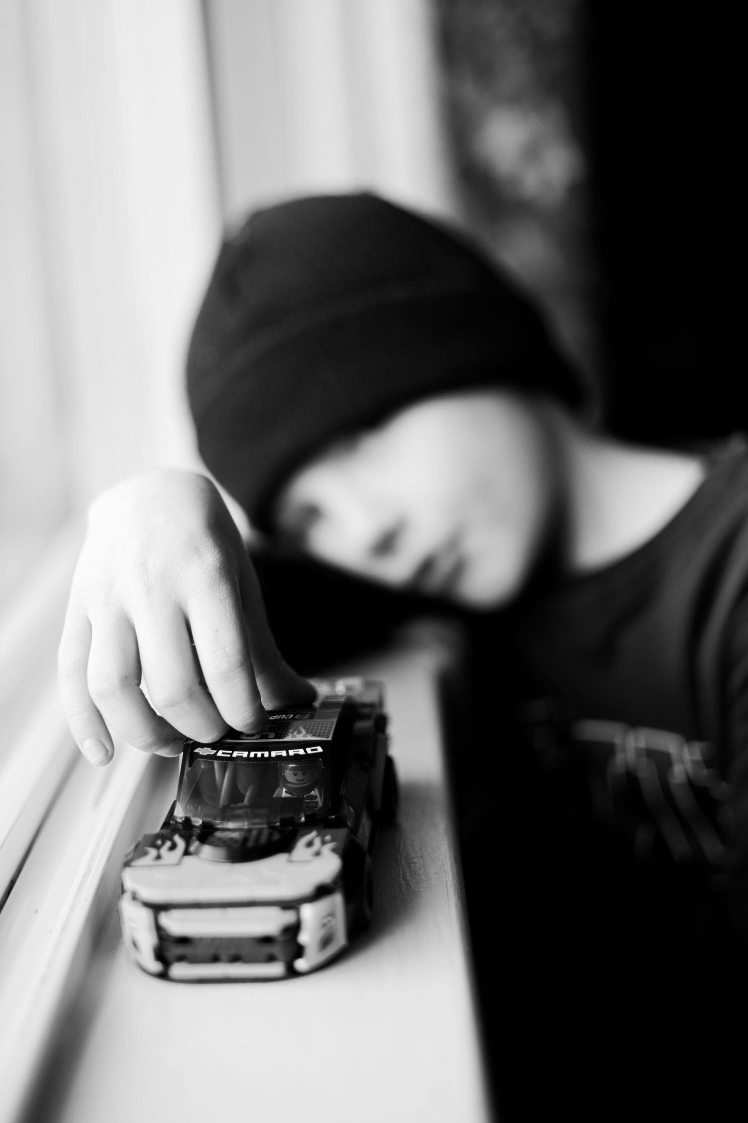 Black and white photo of little boy in beanie playing with a toy car on the window | In-Home Family Photos in Westminster, CO