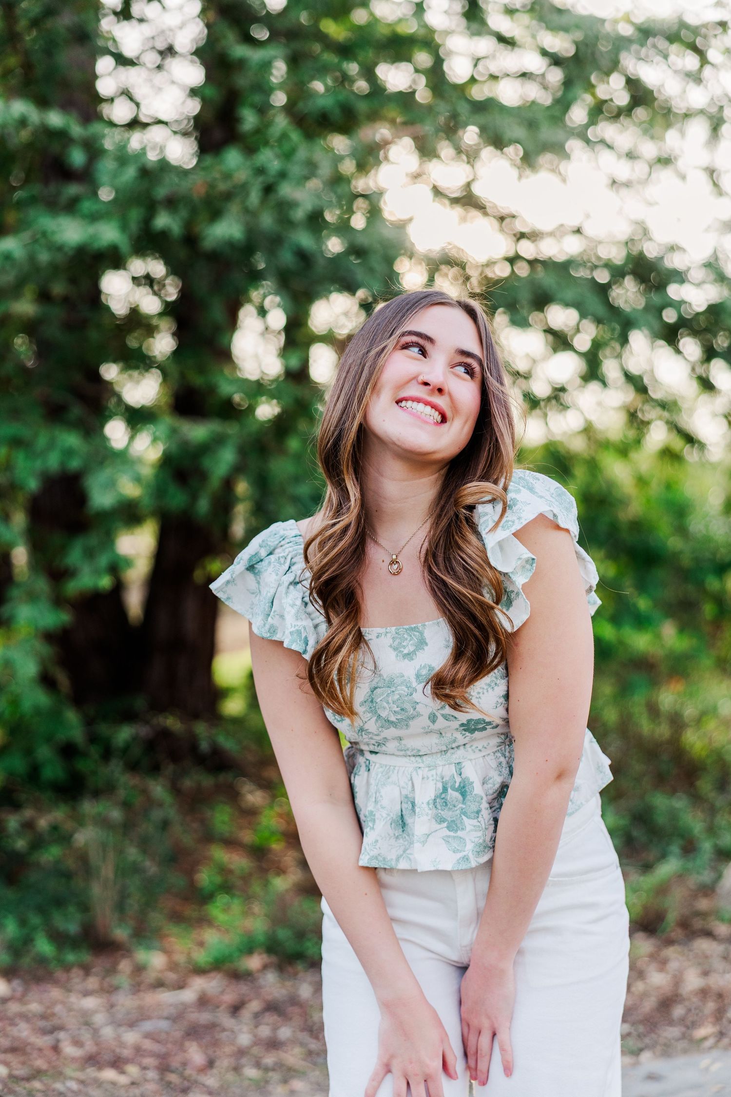 A person in a white dress laughs joyfully while posing outdoors in a sunlit wooded setting.