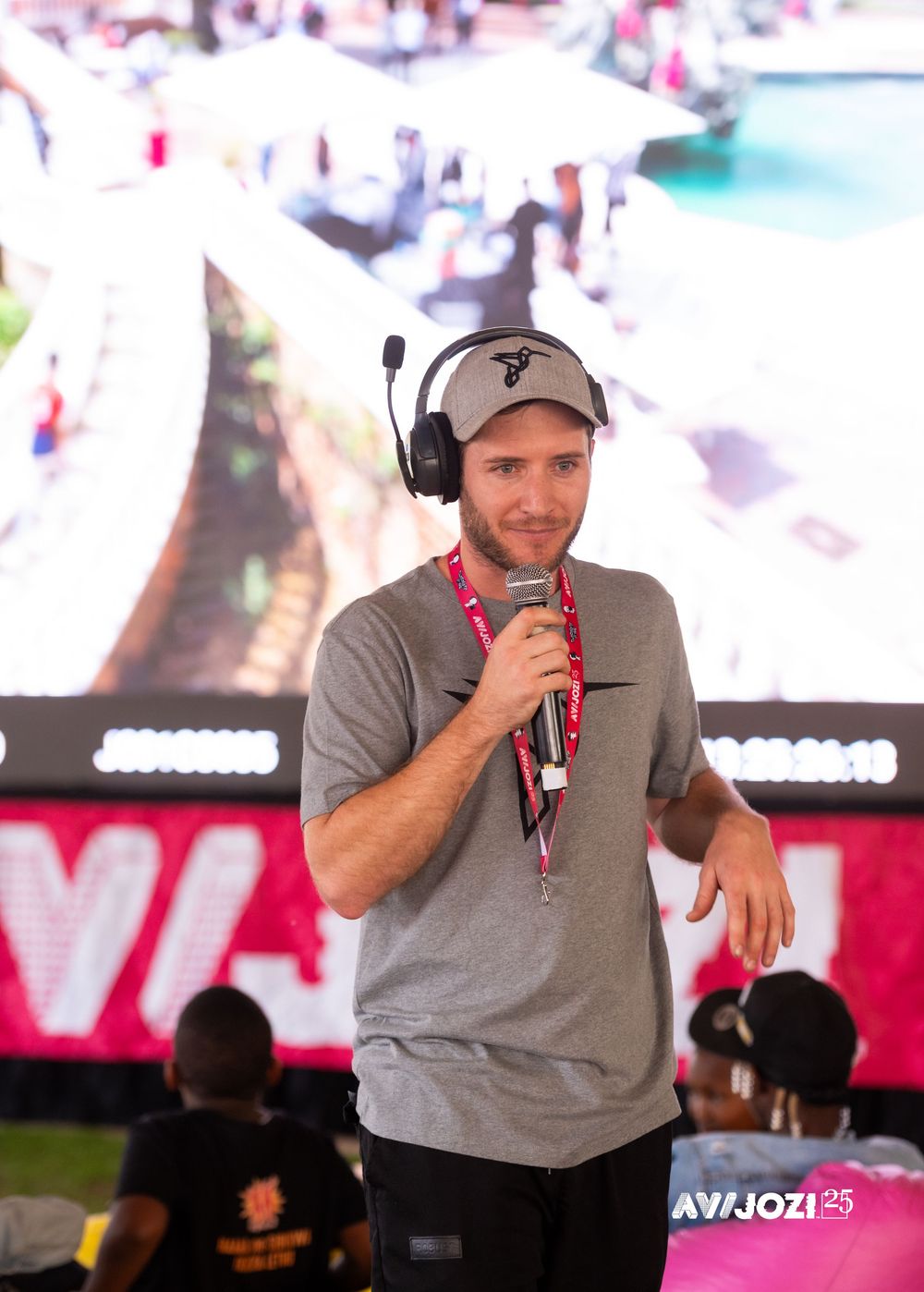 A person in a gray shirt and cap speaks into a microphone on stage with video screens visible in the background.