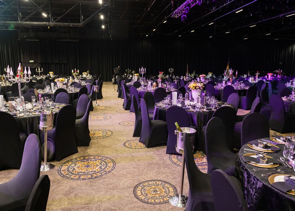 Elegant banquet hall setup with purple-lit tables featuring black chair covers and ornate centerpiece decorations.