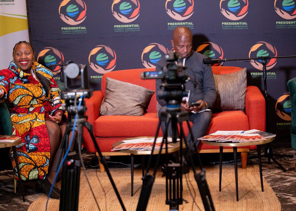 Conference panel setup with orange chairs and video equipment against branded backdrop.