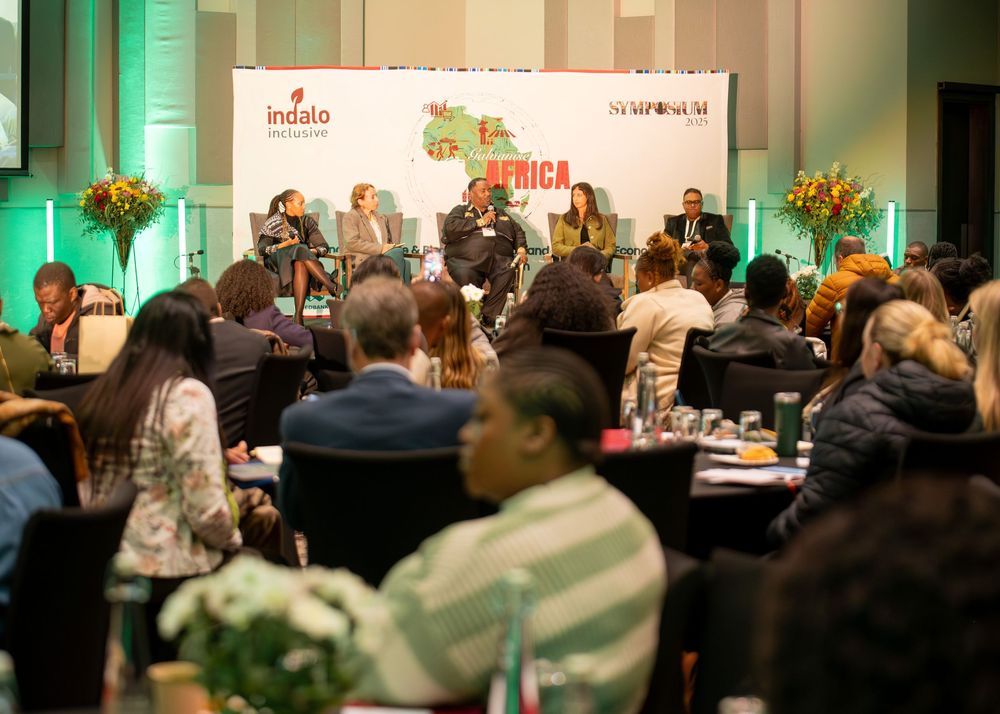 Conference attendees sit in rows at tables during a professional business event with green lighting accents.