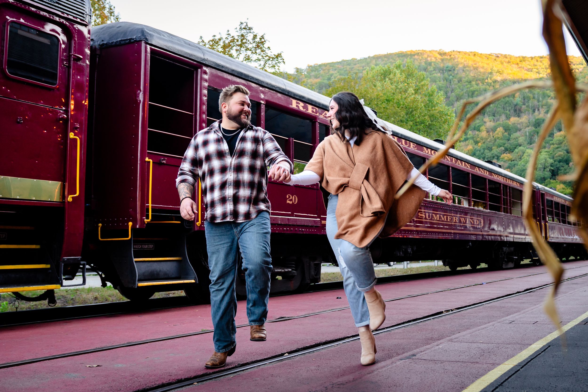 Couple walking along vintage red train cars on scenic mountain railway tracks.