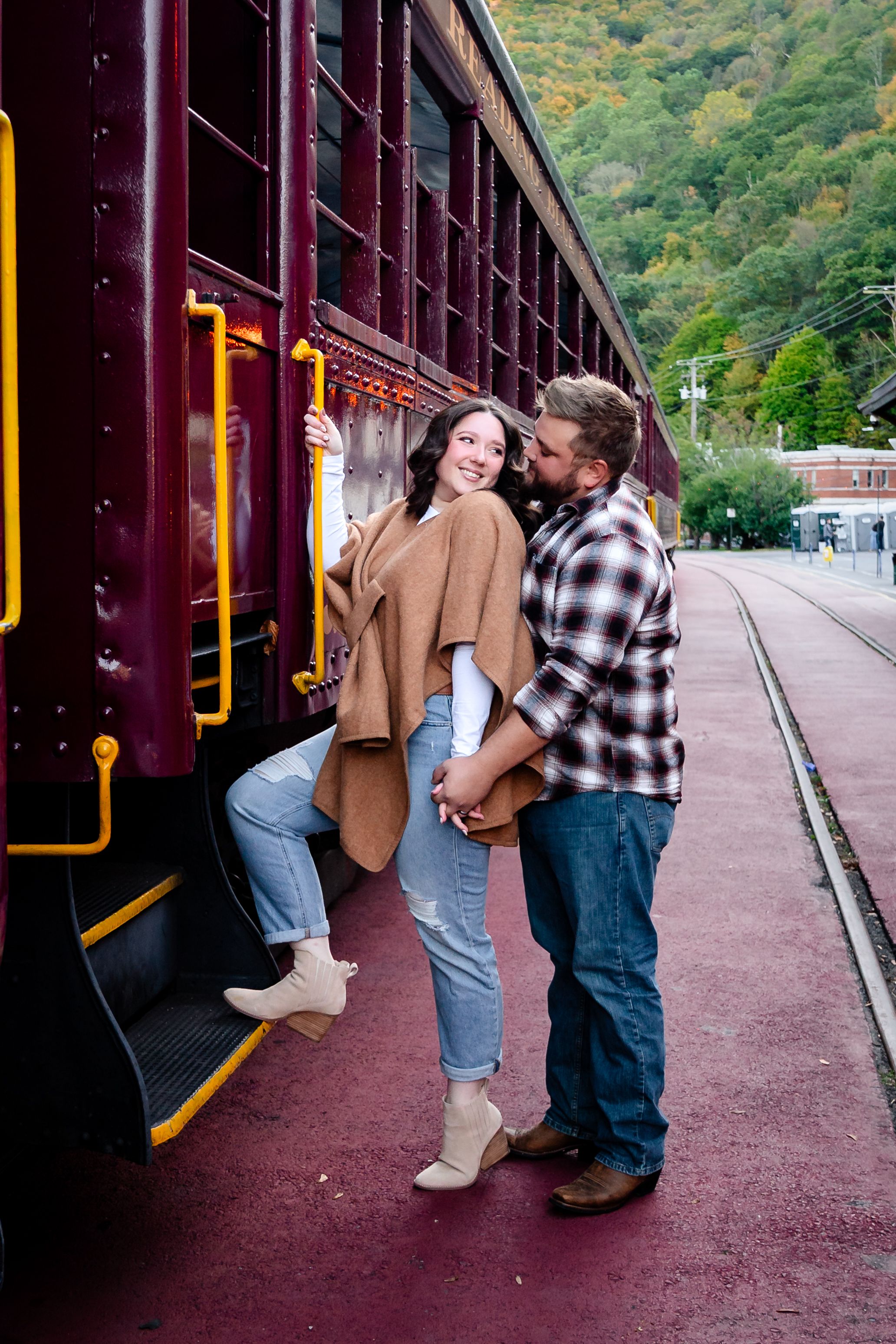 Romantic couple poses by maroon vintage train cars during fall photoshoot.