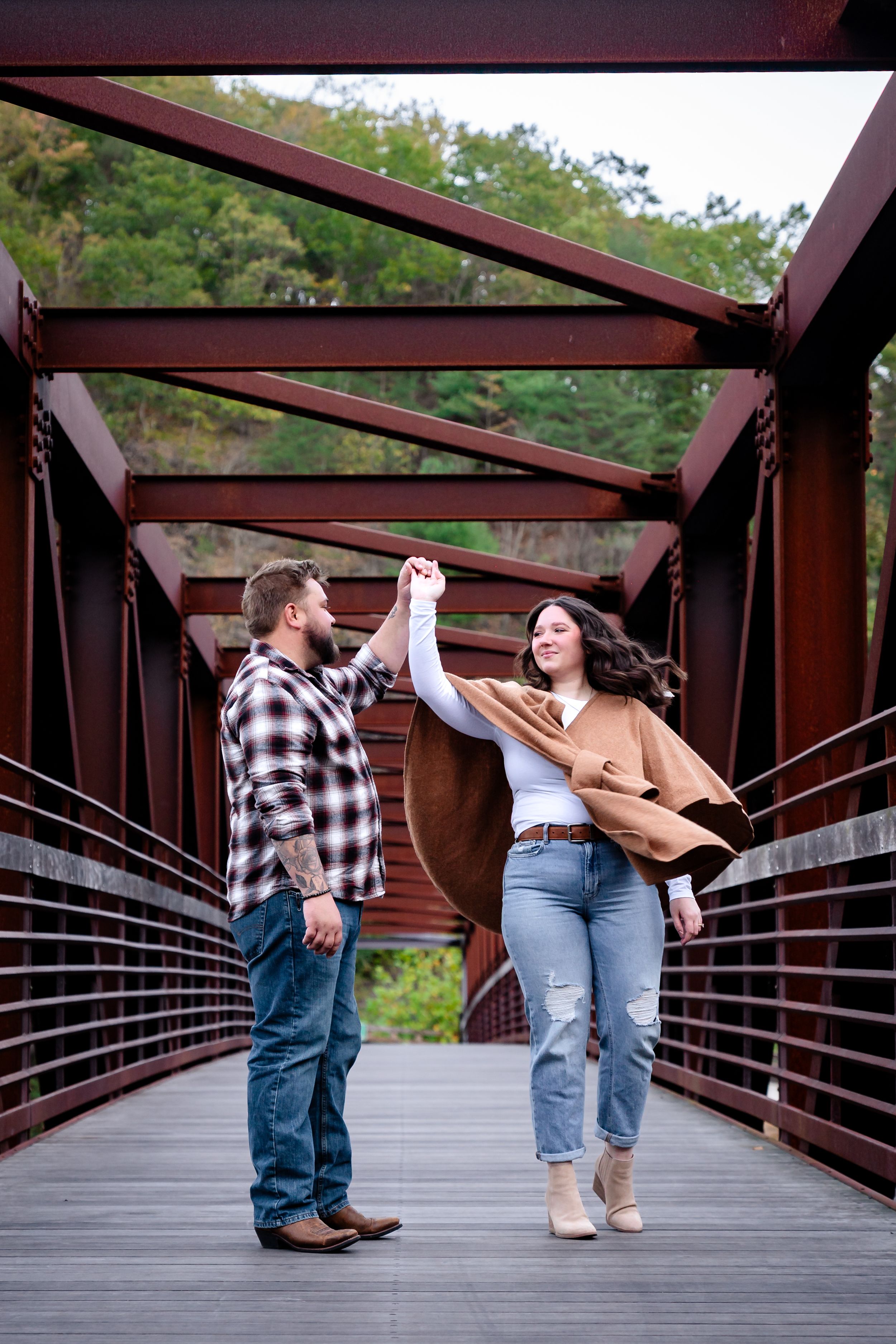 Playful couple dancing and twirling on wooden bridge with metal railings.