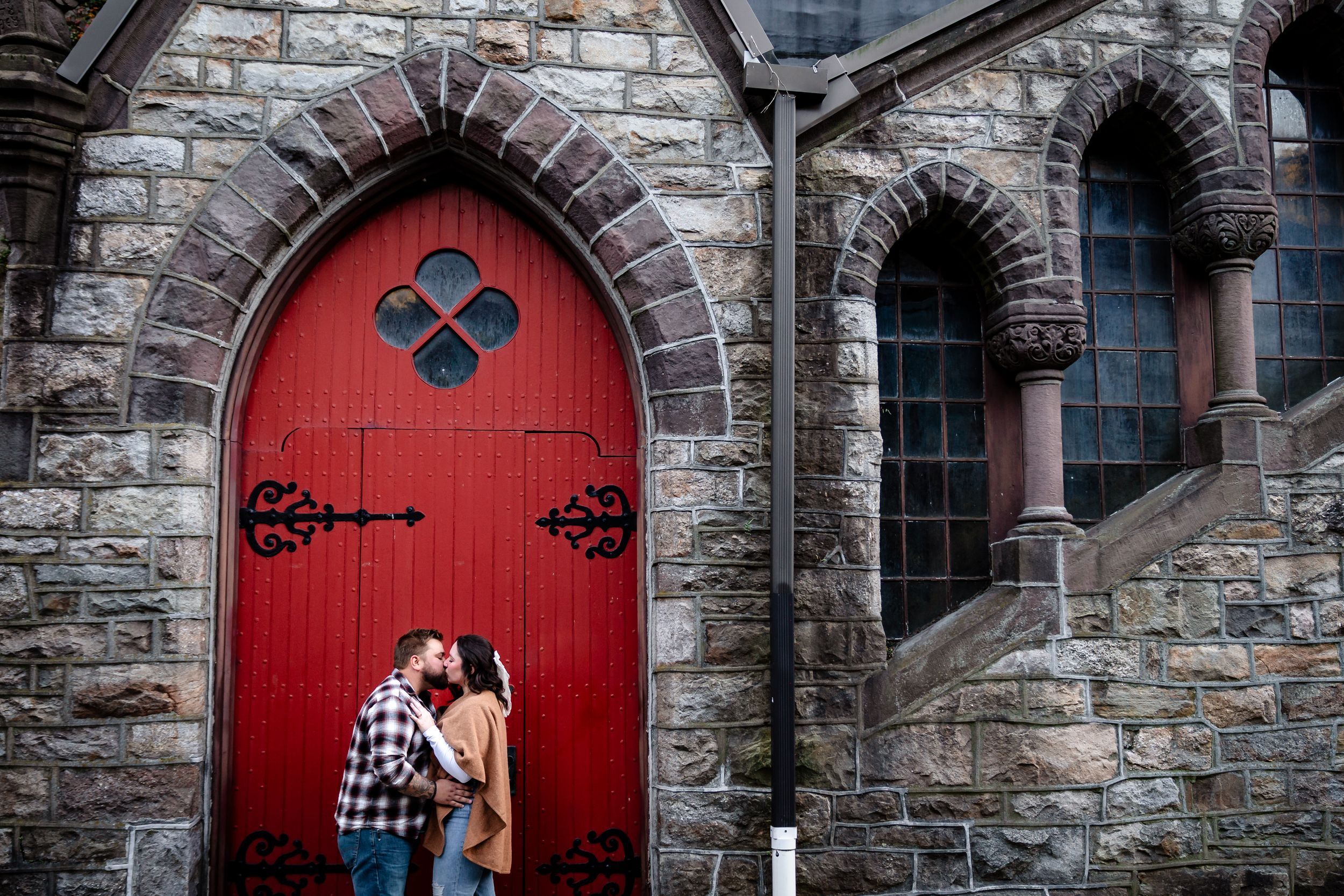 Couple embracing in front of gothic stone church with red doors.