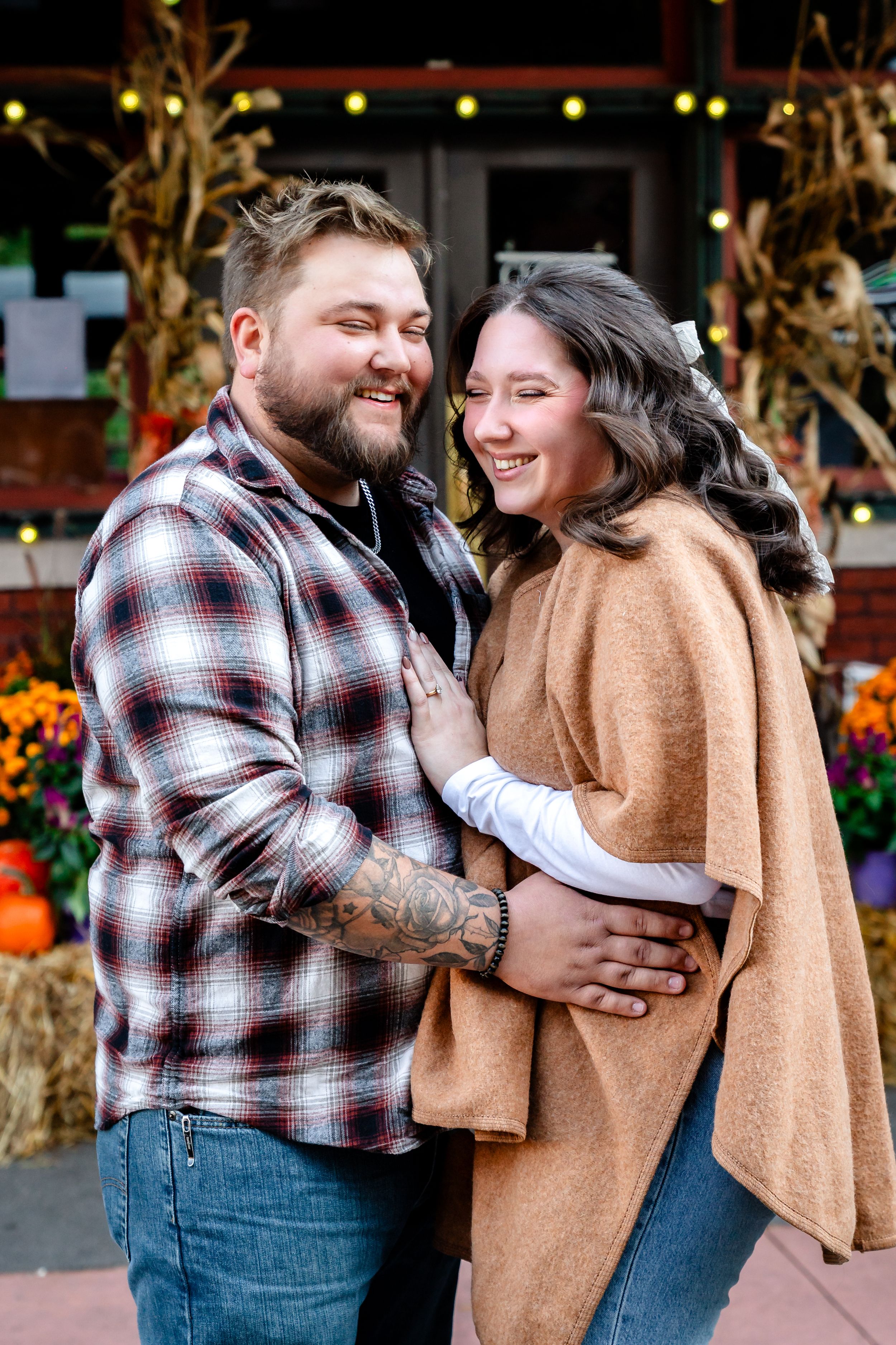 Couple embracing and laughing in front of storefront decorated with fall pumpkins.