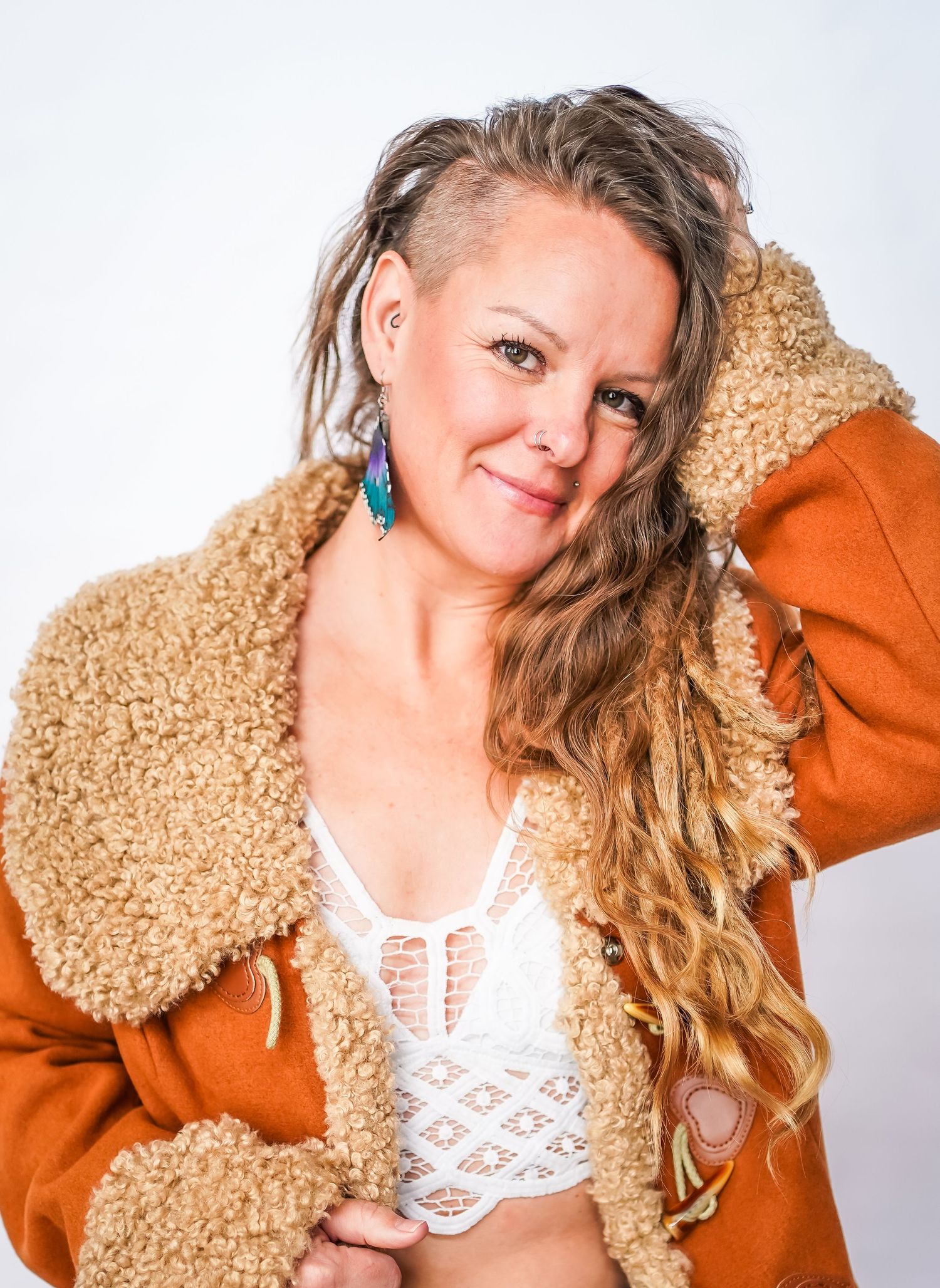 Portrait against white background wearing brown shearling jacket and white lace top with long wavy blonde hair.
