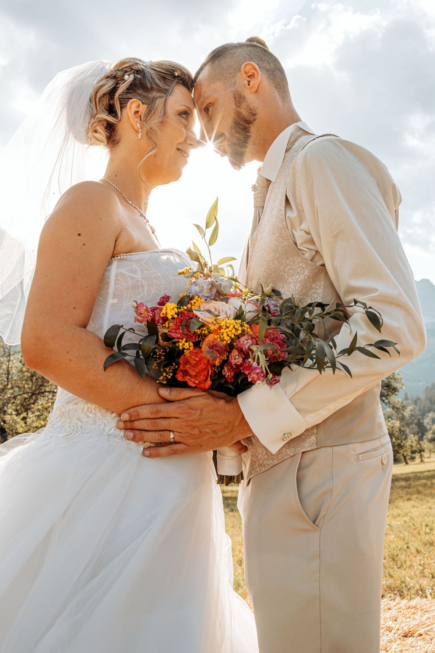 Intimate wedding couple embraces in sunlit field with vibrant orange and red bouquet.