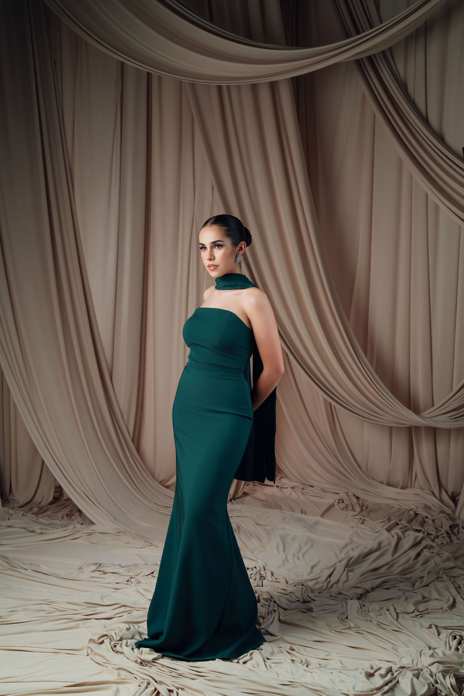 Emerald green formal gown photographed against draped beige fabric backdrop.