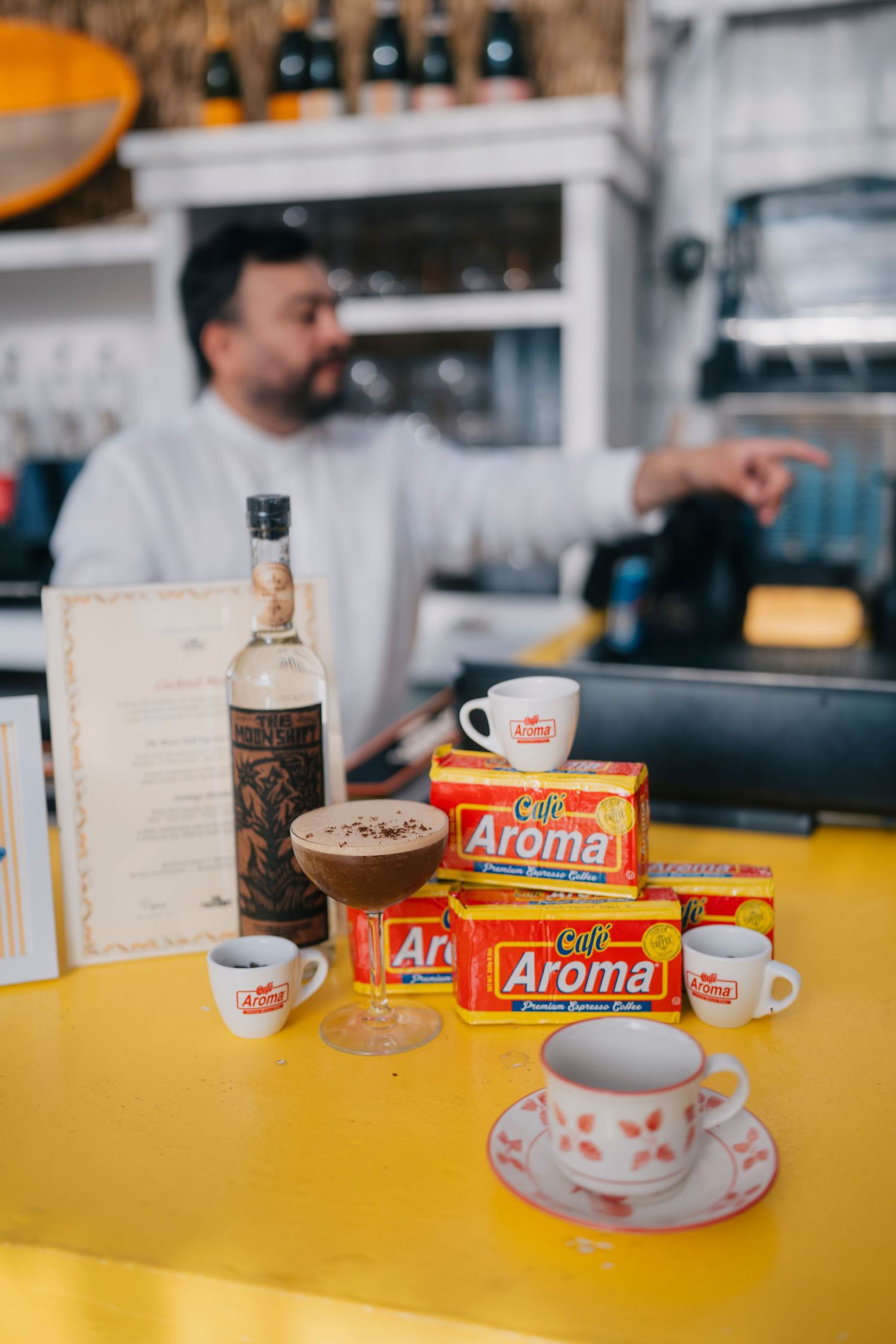 Coffee cups and Aroma brand containers sit on a bright yellow counter in a cafe setting.