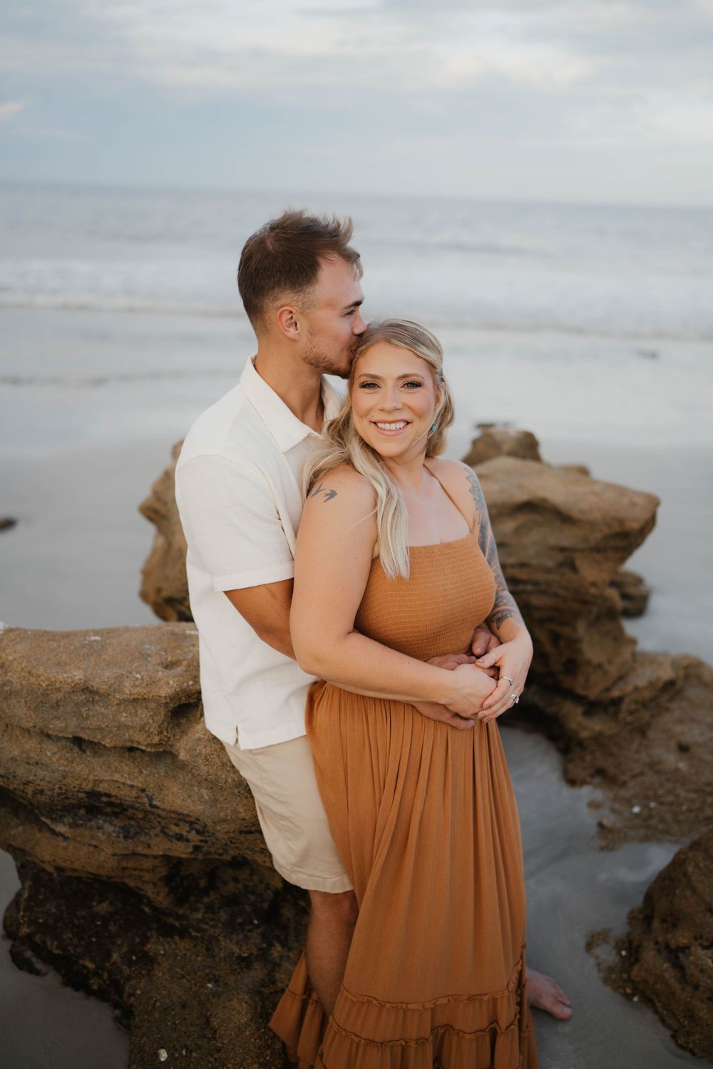 A romantic couple embraces on rocky coastal beach at sunset wearing white and brown outfits.