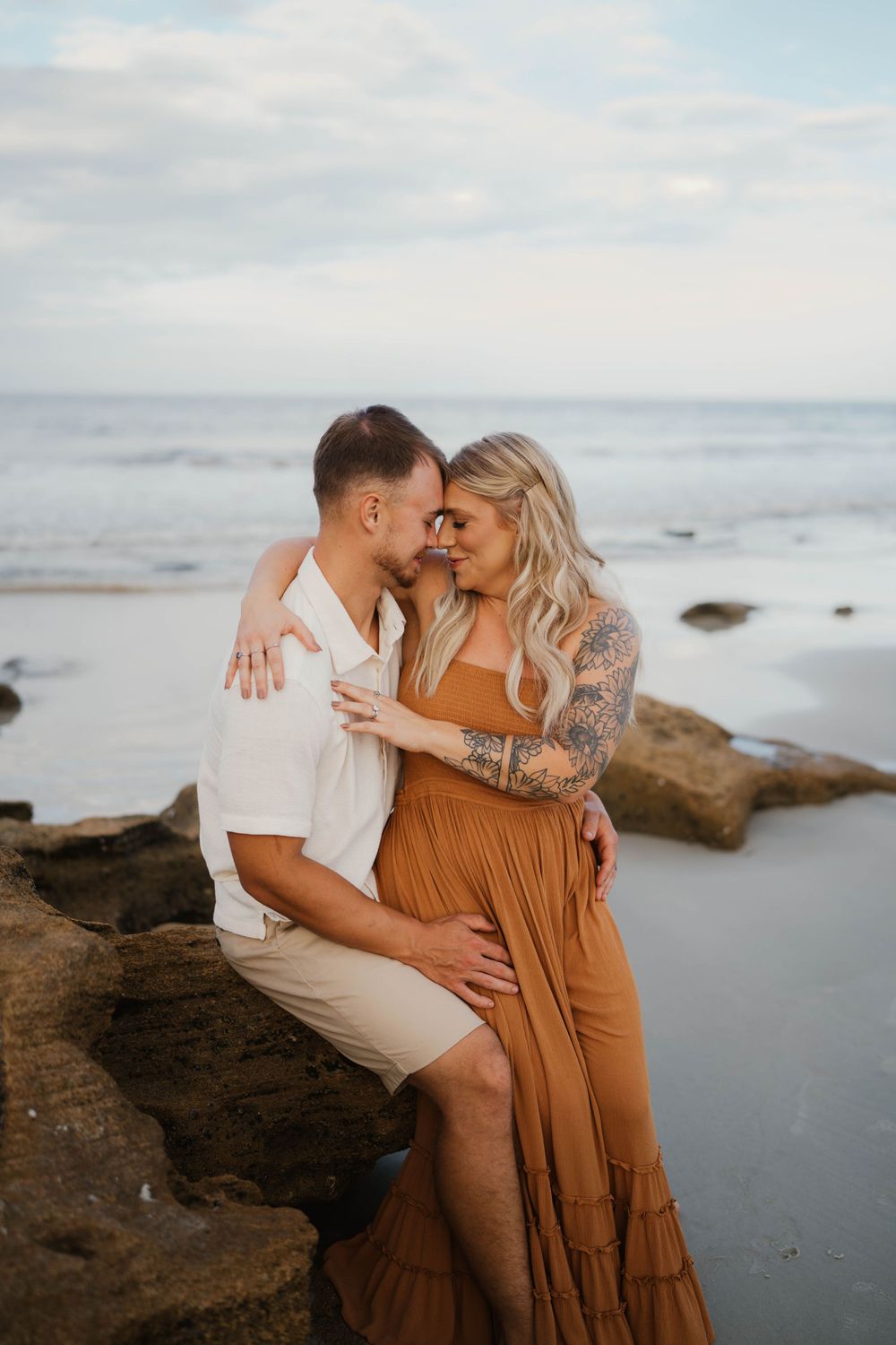 Intimate beachside photo session with couple in coordinated outfits among coastal rocks.