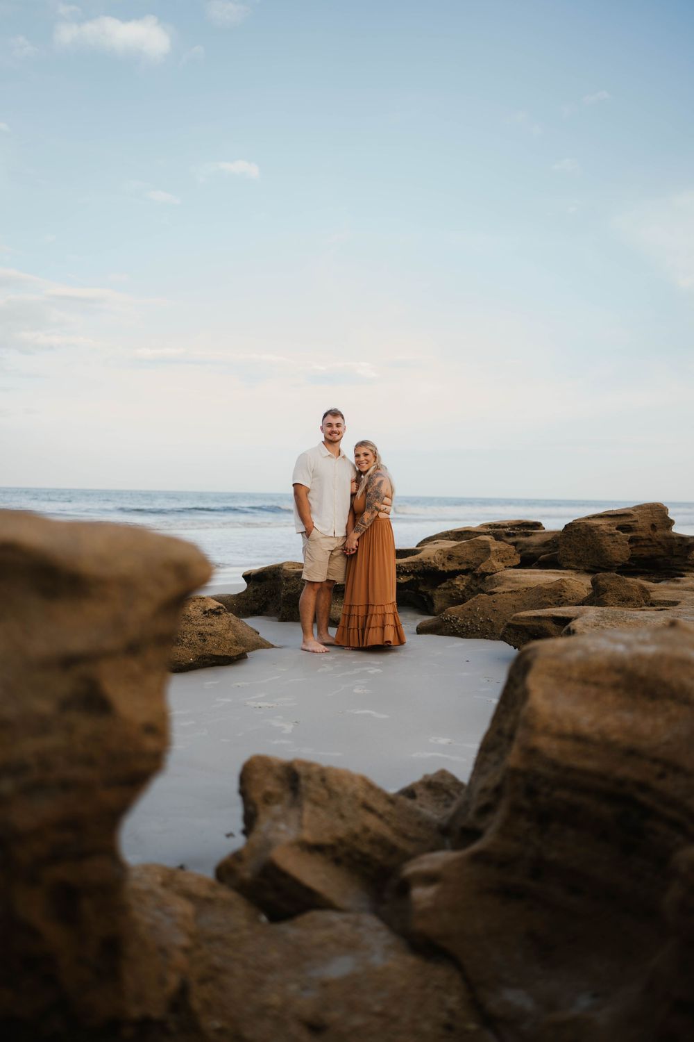 A couple stands together on rocky coastal cliffs at sunset overlooking the ocean.
