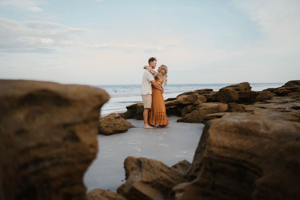 A couple embraces on rocky coastal cliffs at sunset with ocean waves in the background.