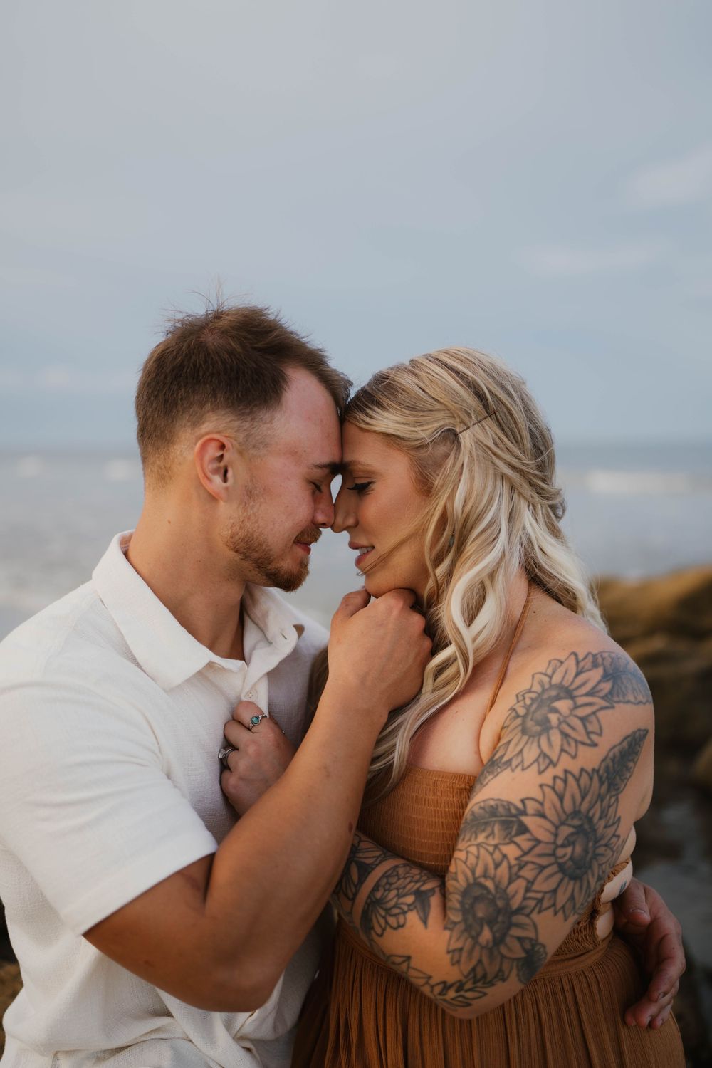 Tender moment captured between couple in white and brown clothing on rocky beach.