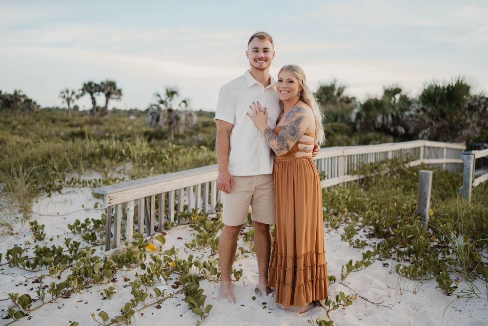 A couple shares a romantic moment on a coastal boardwalk surrounded by sand dunes and beach vegetation at sunset.
