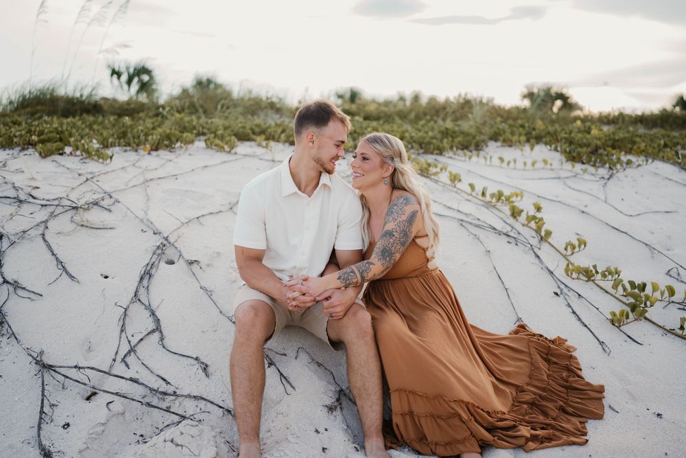 A couple shares a romantic moment on a white sandy beach at sunset with coastal dunes in the background.