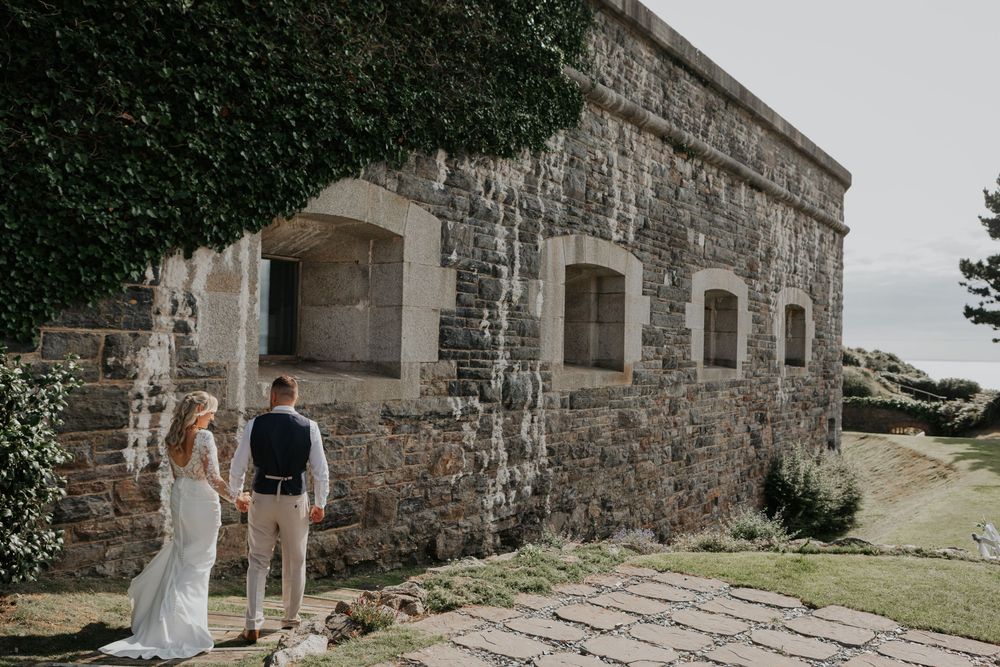 Bride and groom holding hands while admiring a rustic stone building on their wedding day.