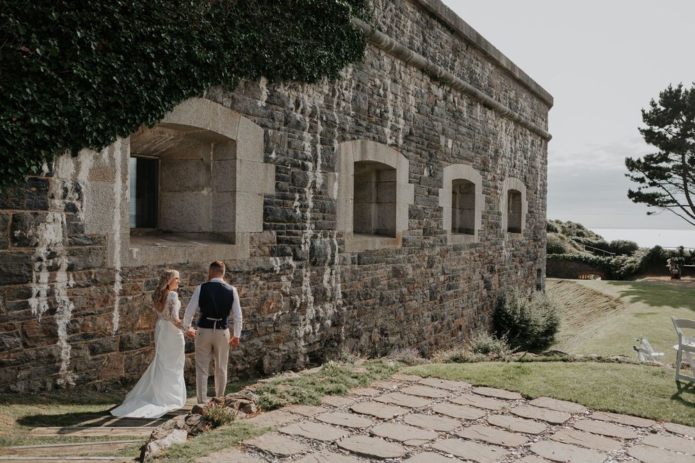Wedding couple standing together beside an ancient stone wall venue on a sunny day.