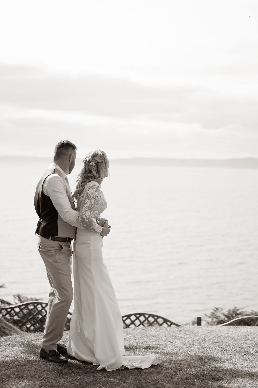Black and white photo of bride and groom embracing with scenic ocean view behind them.