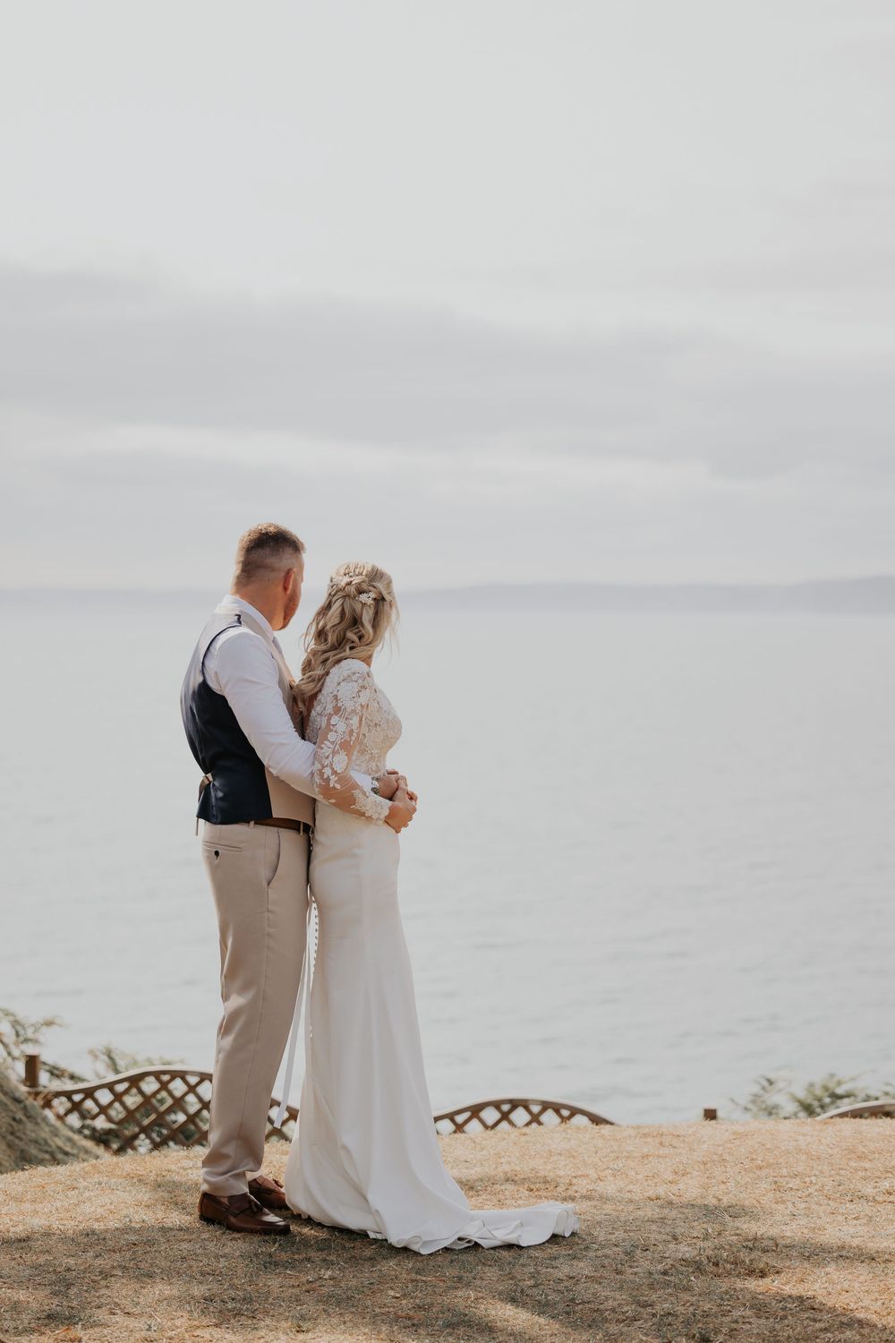 Bride and groom standing together overlooking a calm sea at their outdoor wedding venue.