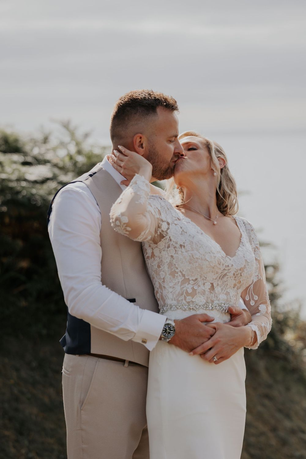 Groom tenderly kissing bride in lace wedding dress with ocean backdrop at golden hour.