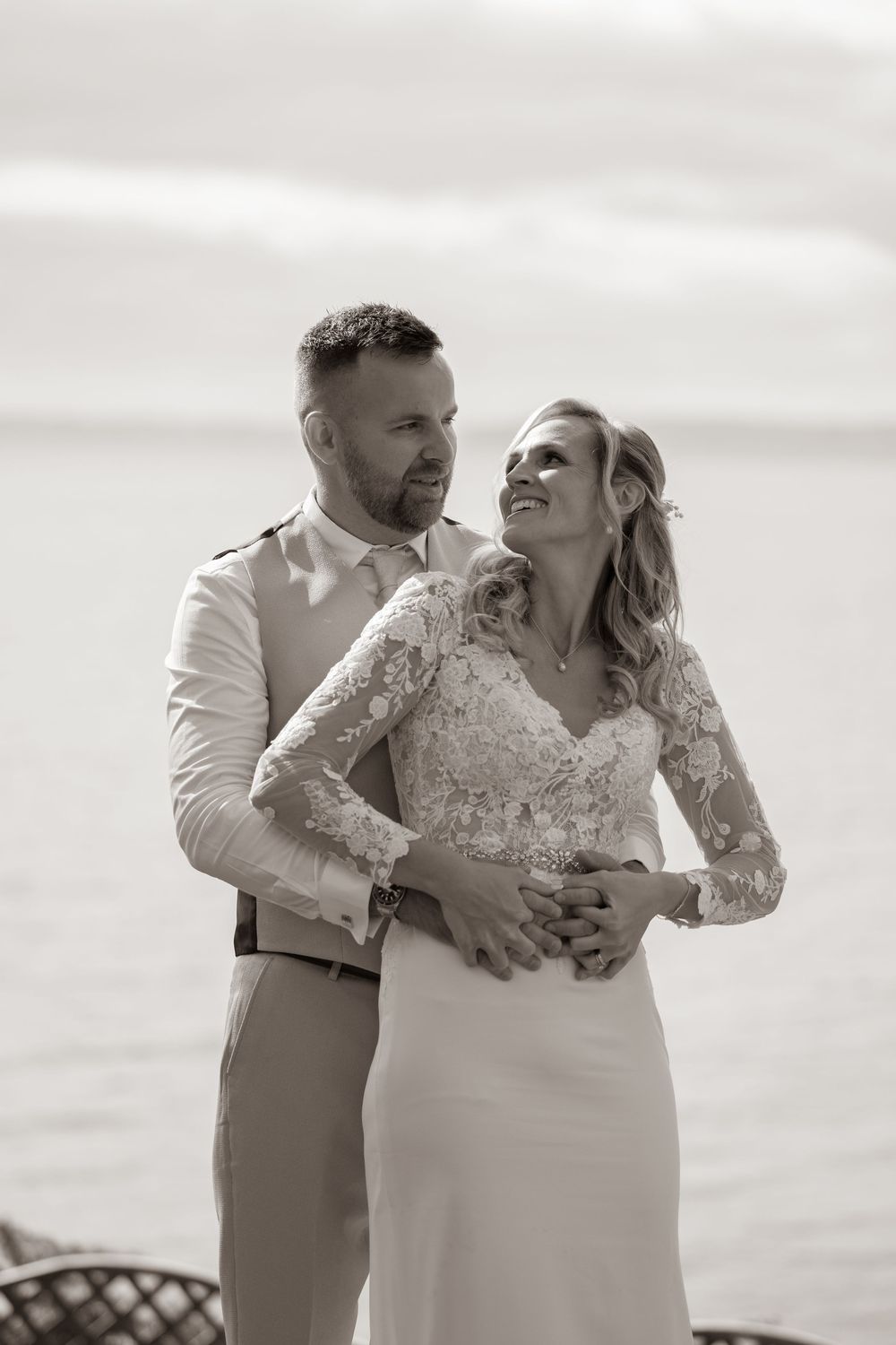 Black and white portrait of smiling bride in lace gown embraced by groom near water.