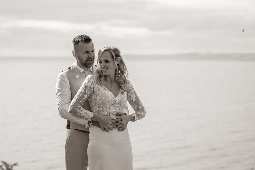 Elegant black and white photo of wedding couple embracing with serene ocean in background.
