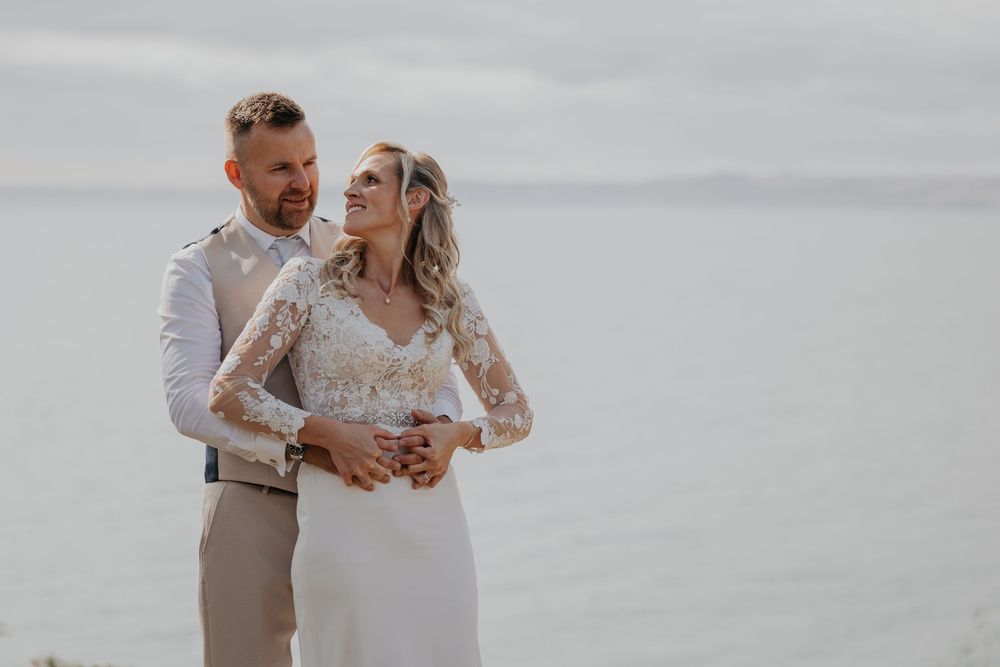 Bride in long-sleeve lace gown smiling up at groom with calm sea behind them.
