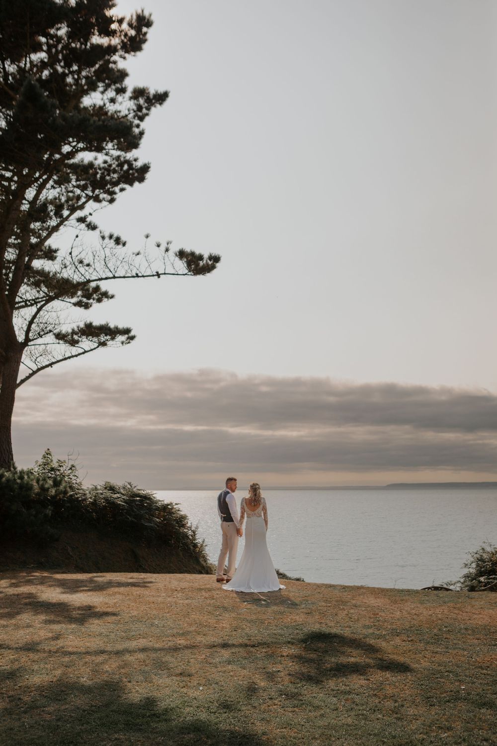 Bride and groom holding hands on grassy cliff overlooking a dramatic coastal sunset view.