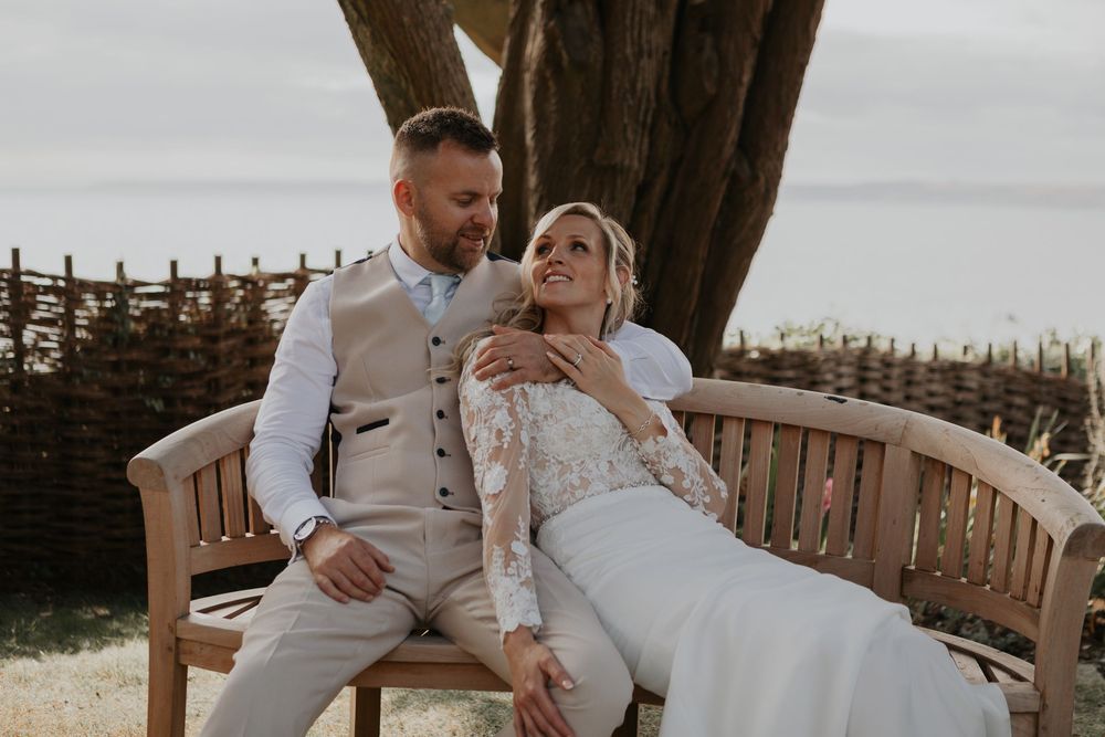 Bride reclining on wooden bench with groom beside a wicker fence and large tree.