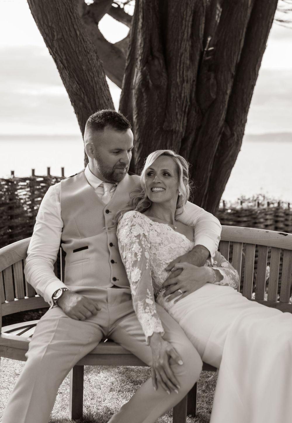 Bride and groom sitting intimately on a wooden bench with ocean views in a black and white wedding portrait.
