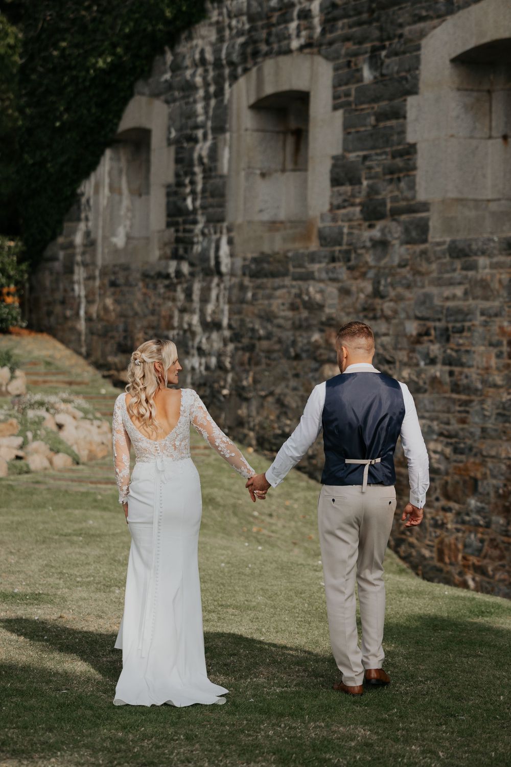 Bride in lace gown and groom holding hands walking past a historic stone wall at wedding venue.