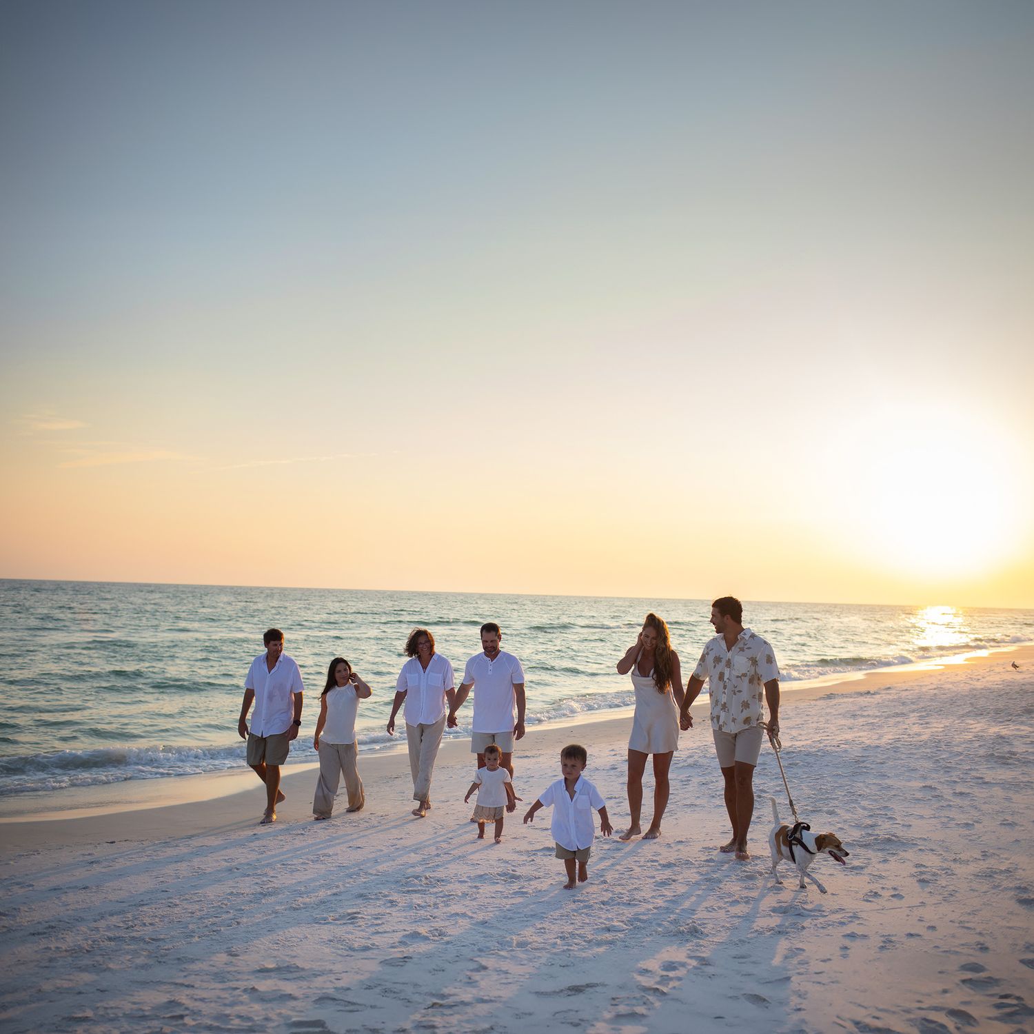 Family group walking along white sandy beach at sunset with ocean waves and golden light in the background.
