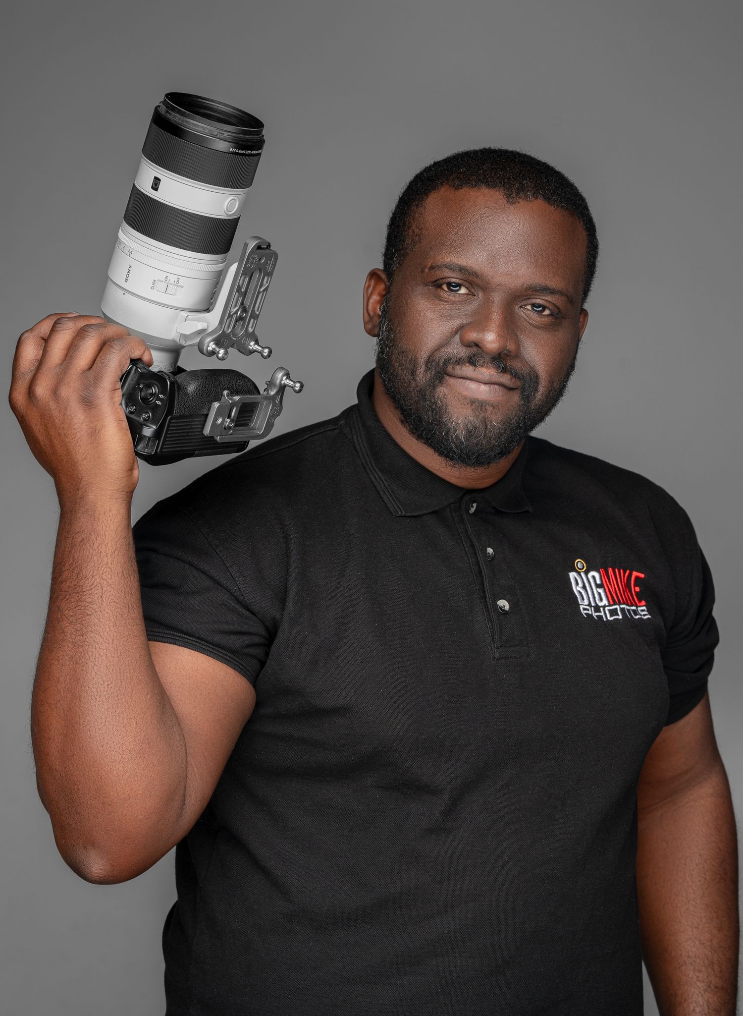 Professional photographer in black polo shirt holds camera lens while posing against gray studio background.