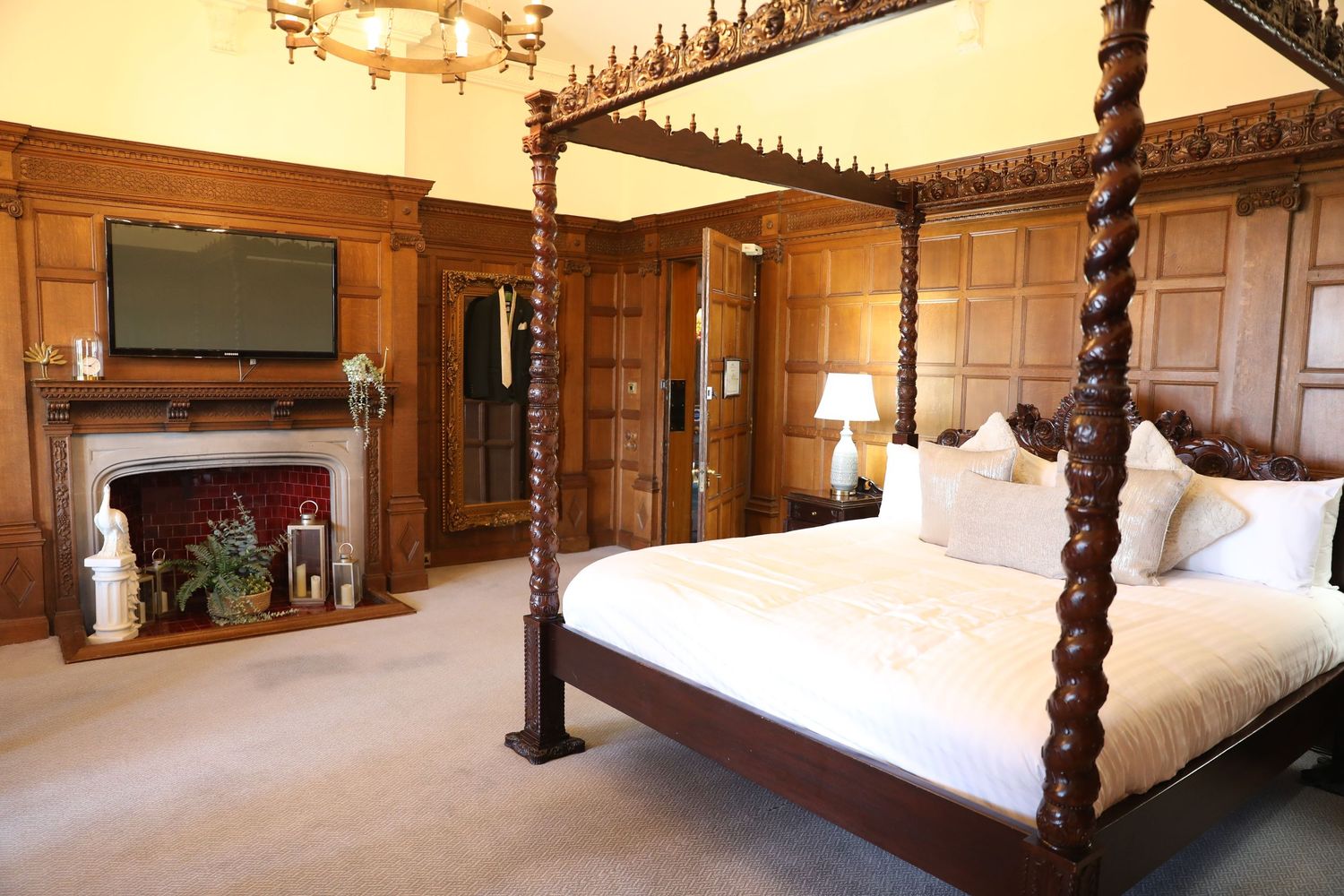 A luxurious four-poster bed with ornate wooden carvings sits in an elegant historic bedroom with rich wood paneling.