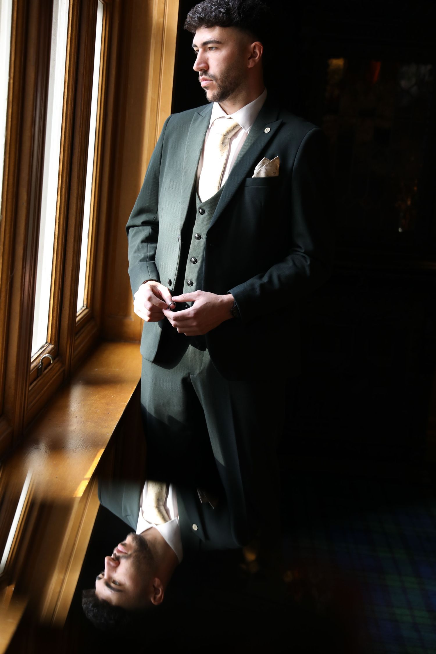 Person in formal attire stands by sunlit windows in a dark hallway creating dramatic lighting and shadows.