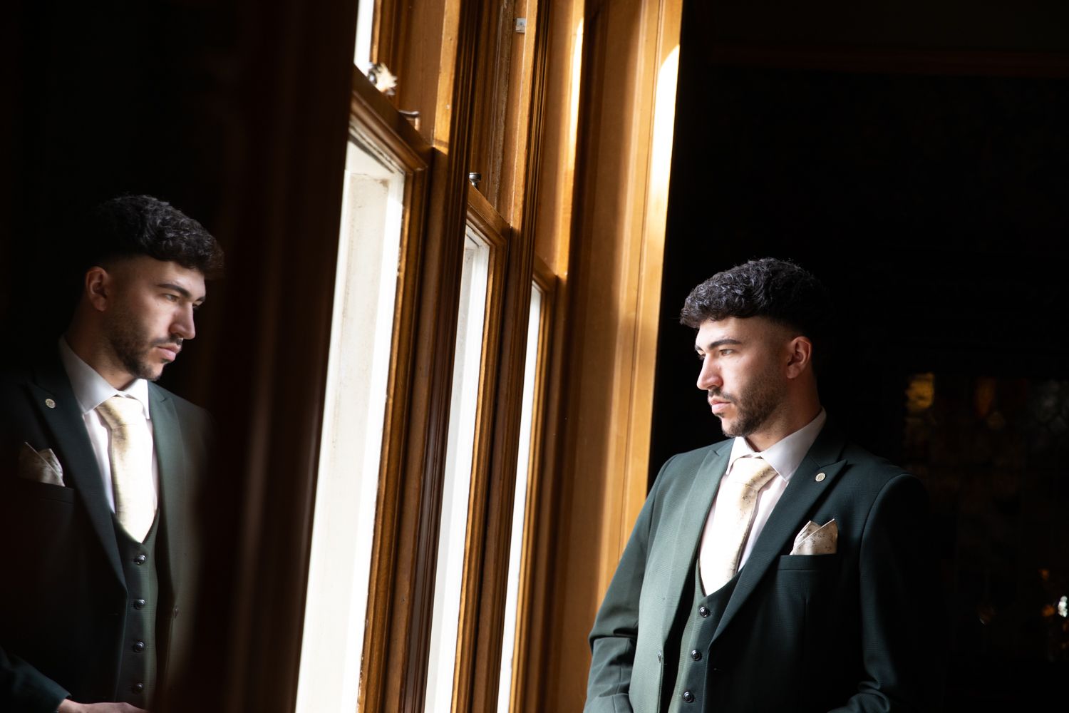A person in a suit jacket stands in dramatic lighting near wooden window frames creating a moody portrait effect.