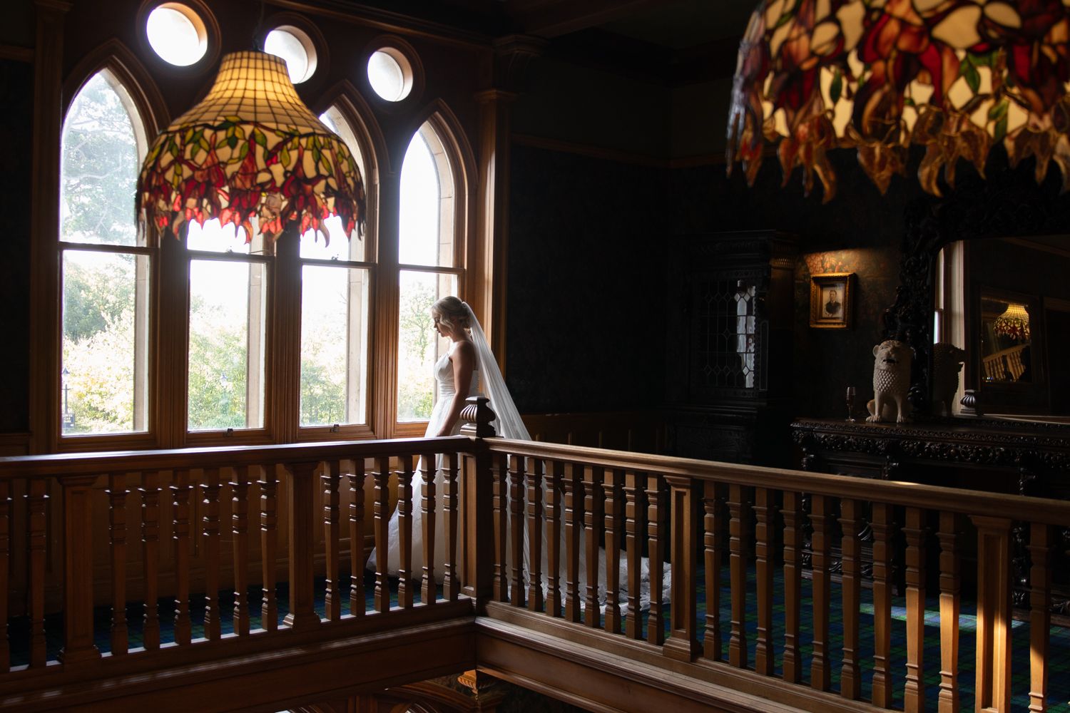 Elegant dark wood staircase and balustrade in a historic mansion with ornate lighting fixtures and stained glass windows.
