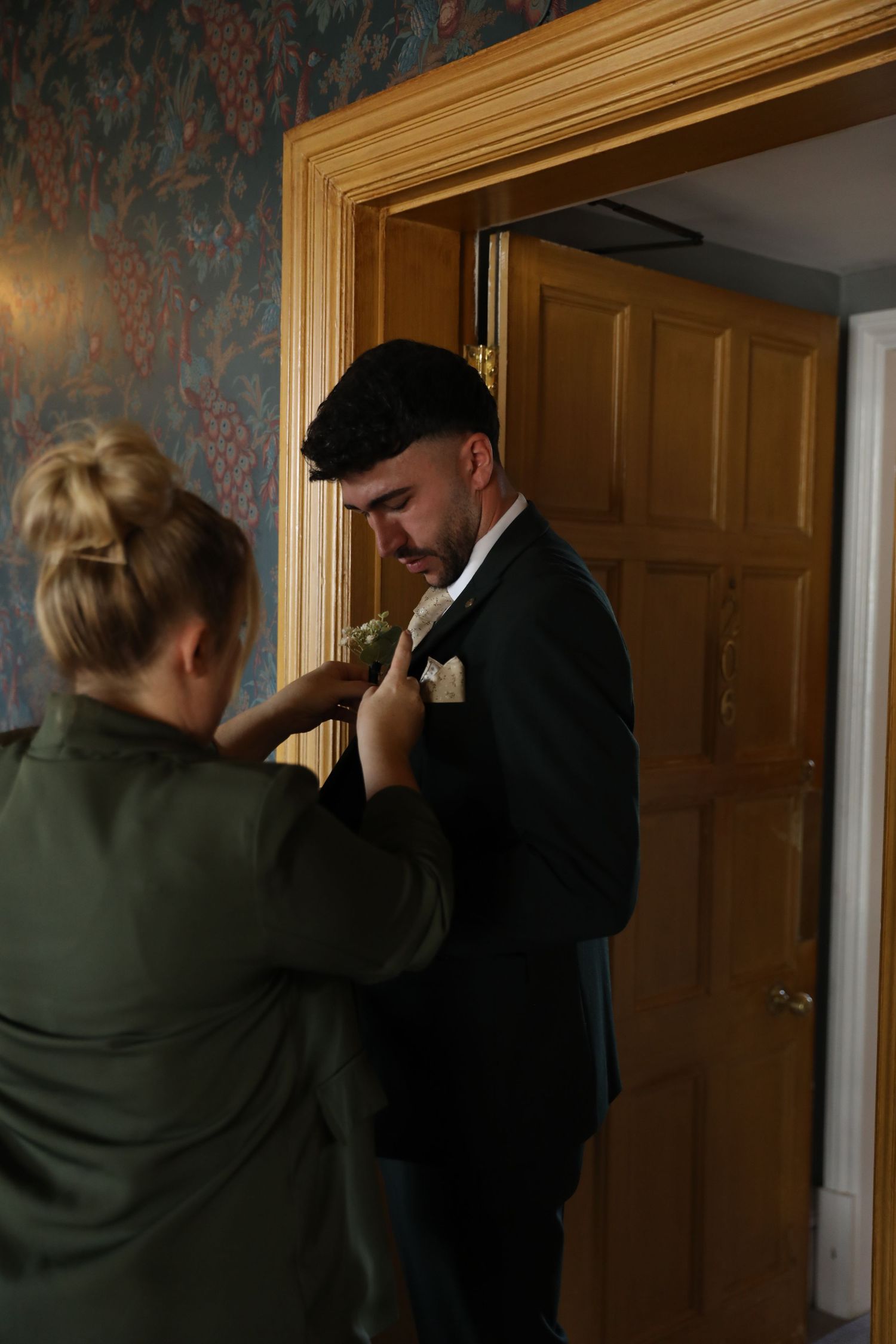 Someone helps adjust a boutonniere on a formal black suit jacket during wedding preparations.
