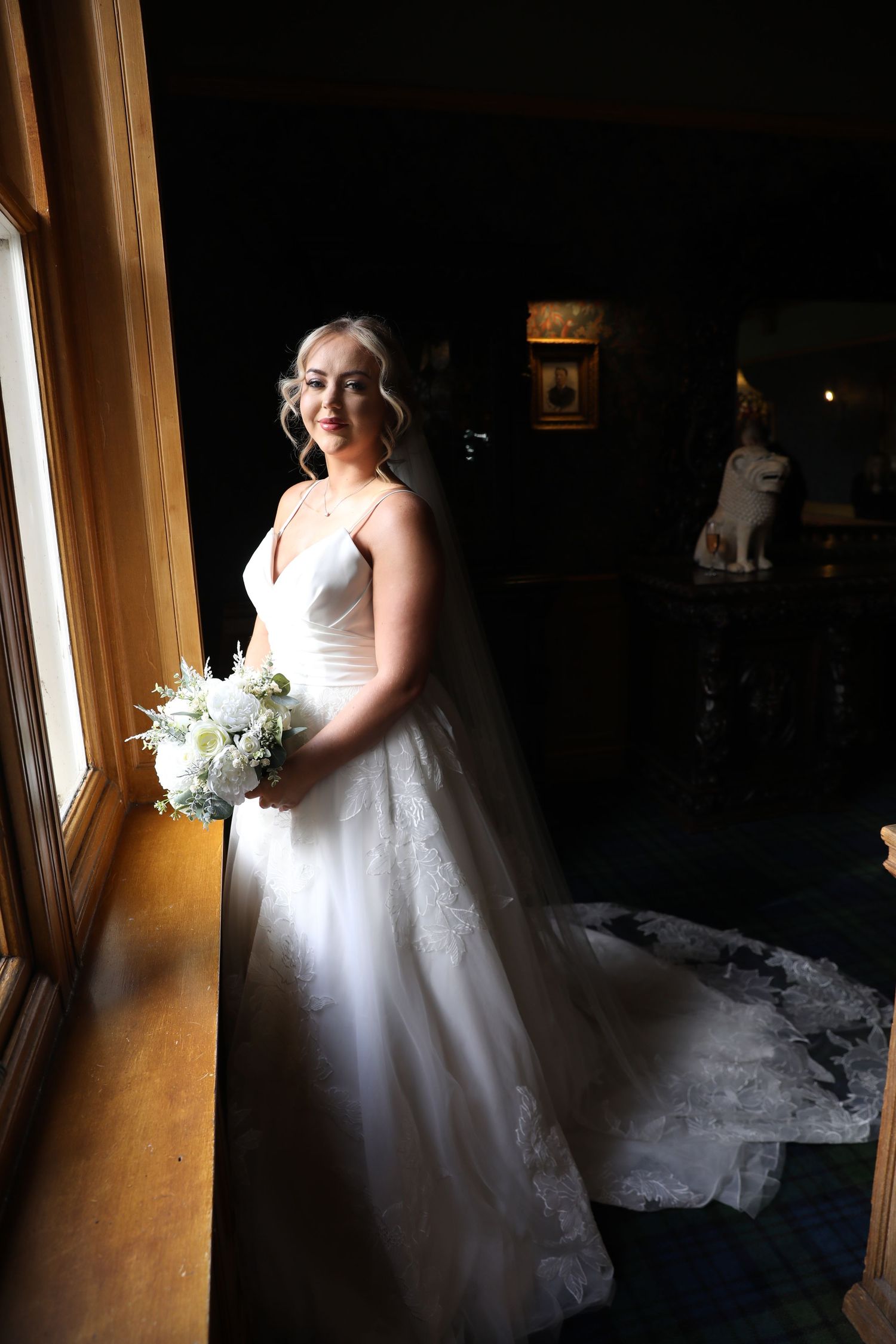 A bride in a white wedding dress stands near a window creating a dramatic silhouette effect in natural light.