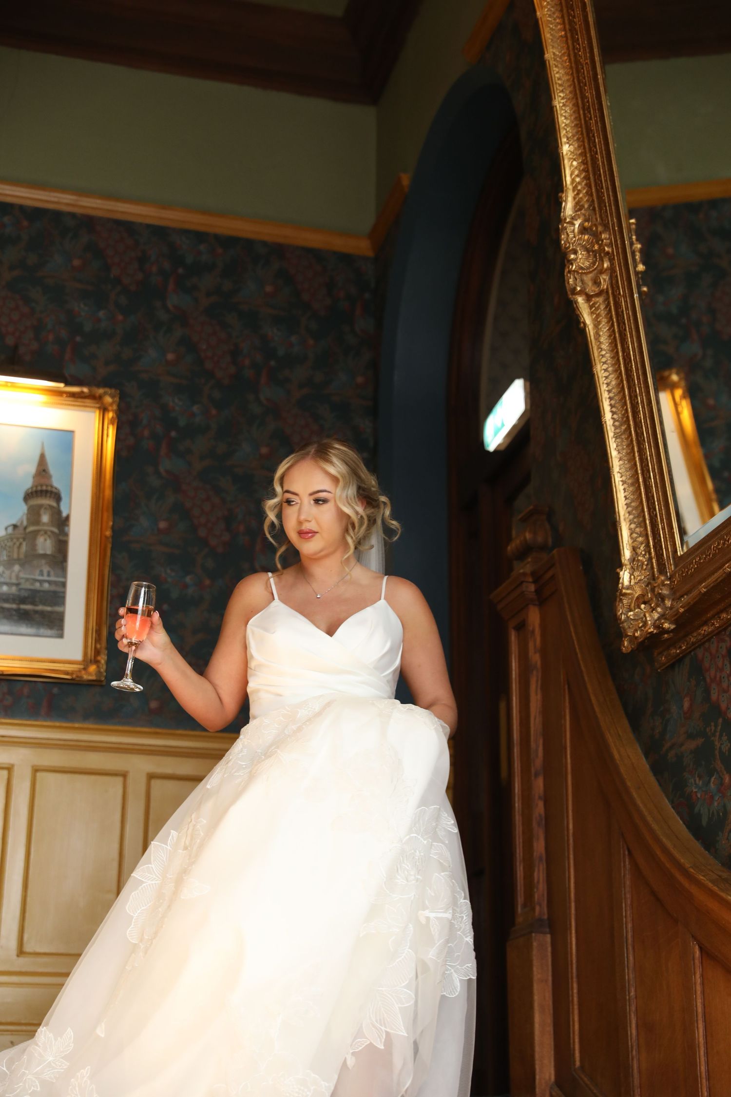 A bride in a white lace wedding dress poses on an elegant staircase with ornate wallpaper and gold-framed mirrors.