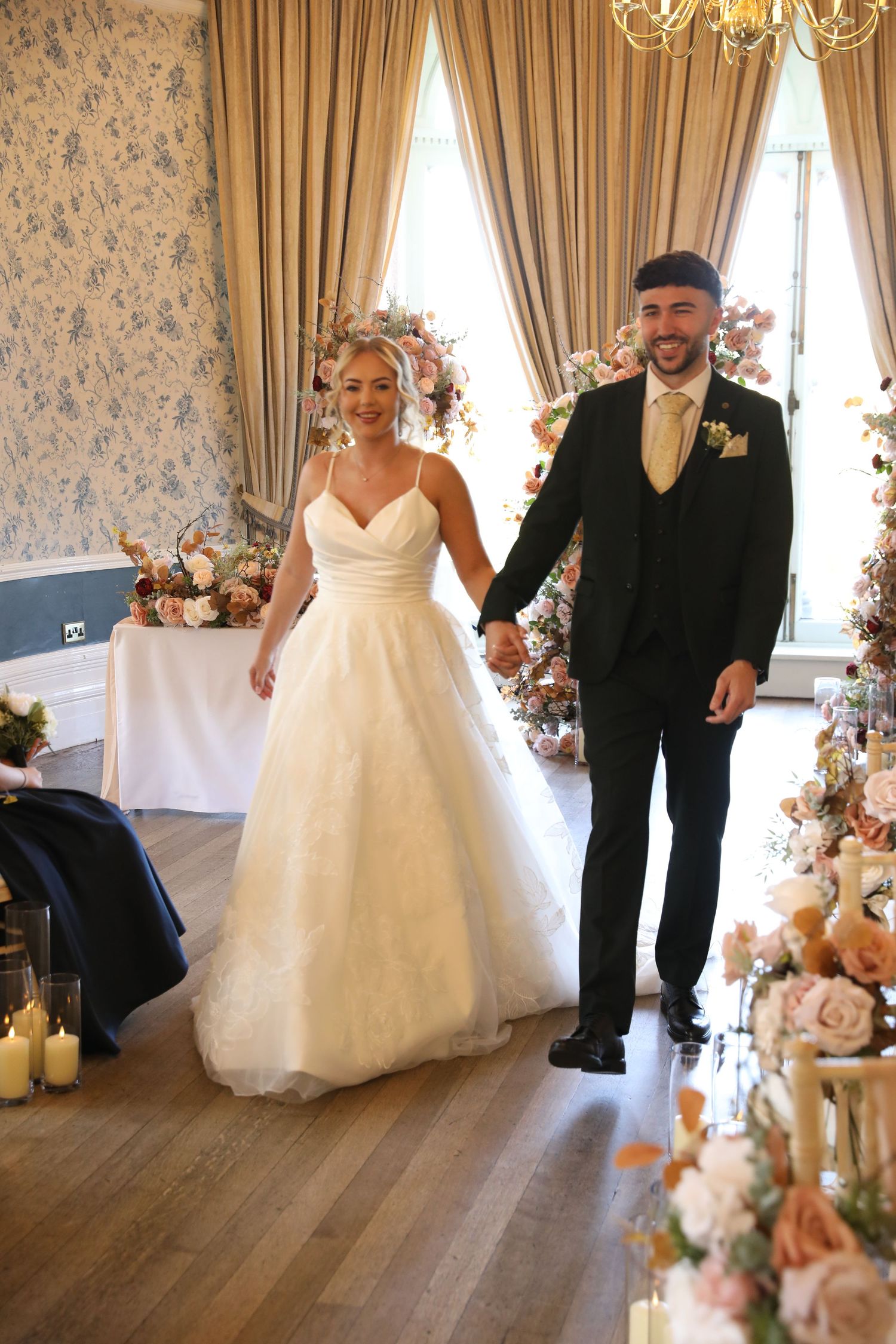 A wedding couple walks together in an elegant room with floral decorations and cream-colored curtains.