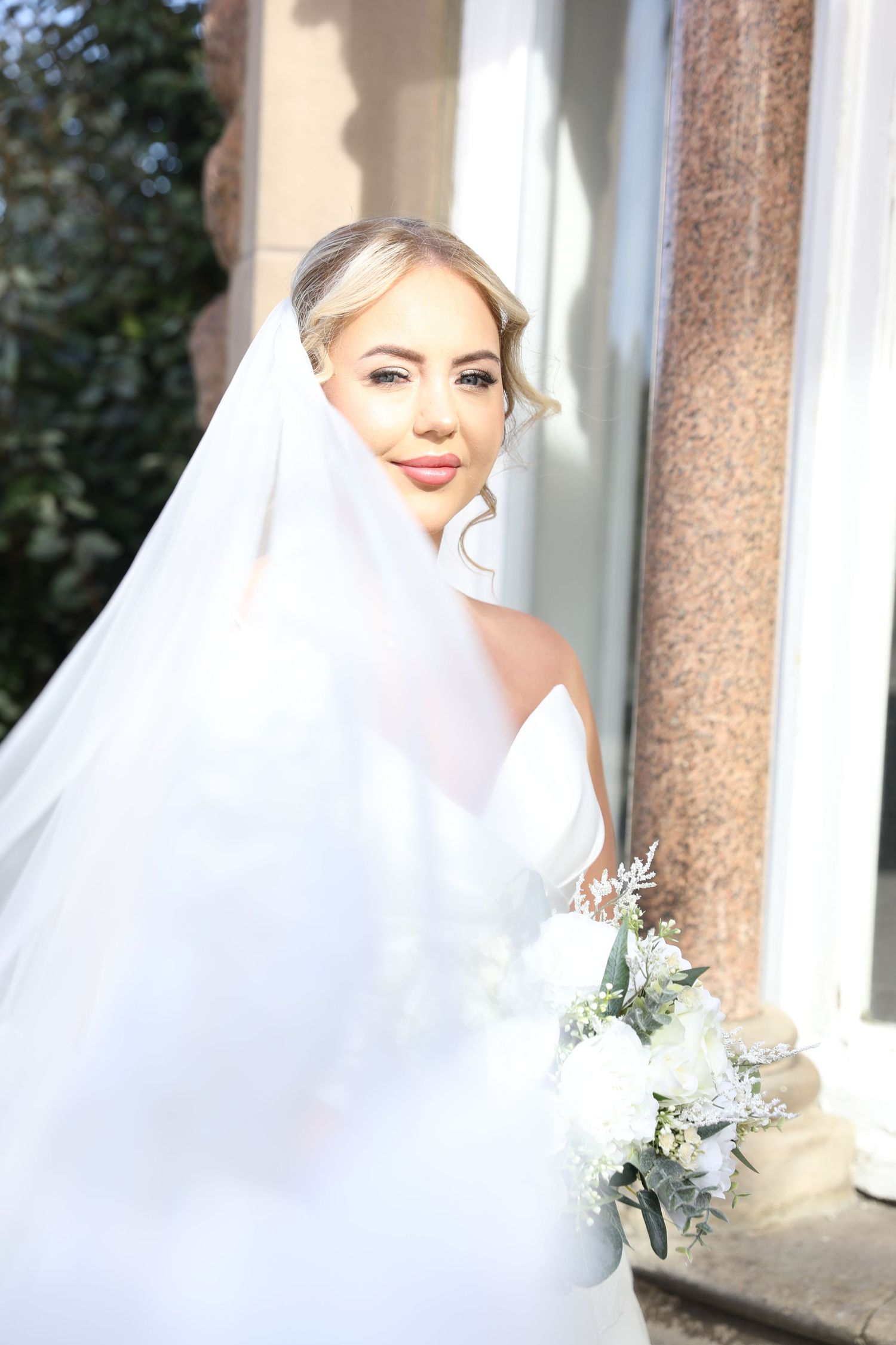 A bride in a white wedding veil poses elegantly against white columns in an outdoor setting.